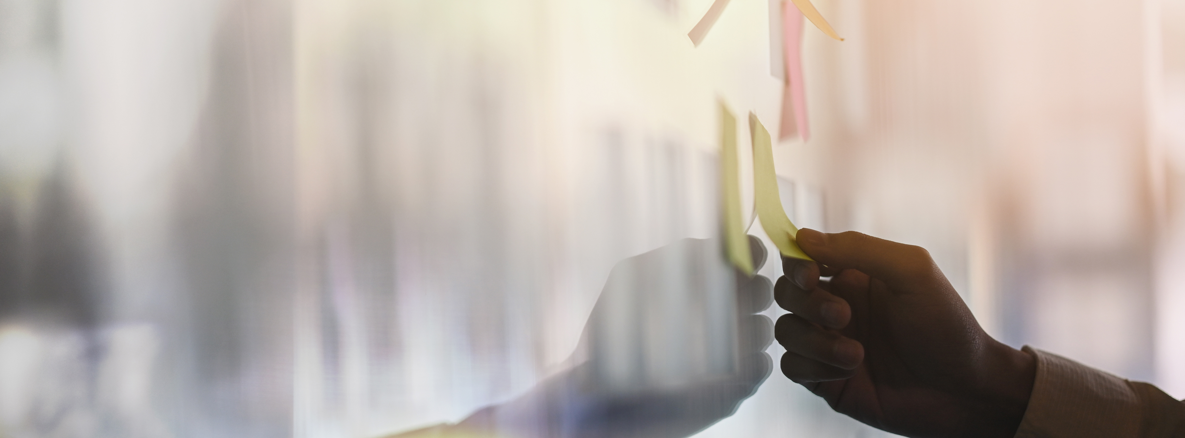 Eine Hand platziert einen gelben Haftnotizzettel an einer glatten Oberfläche. Hintergrund und andere Elemente sind unscharf, der Fokus liegt auf dem Handeln mit den Notizen.