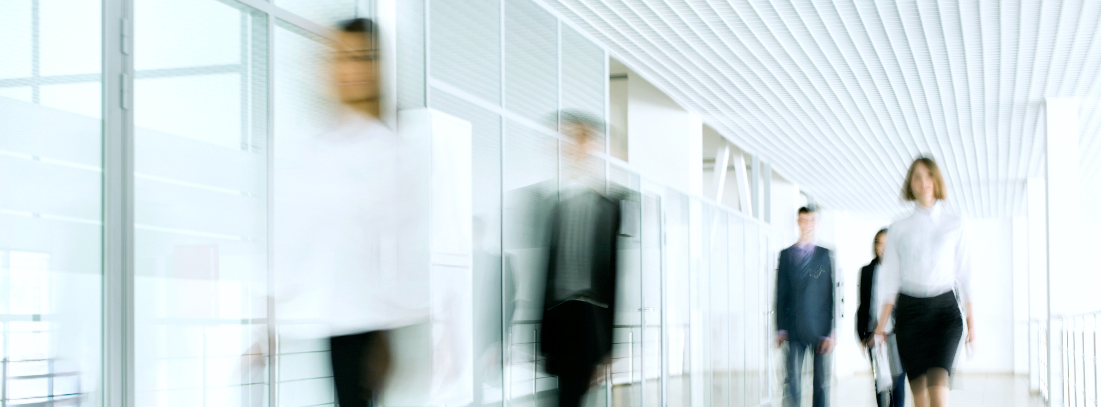 Eine Gruppe von Menschen in formeller Kleidung geht in einem modernen Büroraum. Die Personen sind leicht verschwommen und vermitteln eine dynamische Atmosphäre. Es herrscht ein heller und offener Raum mit großen Fenstern und minimalistischem Design.
