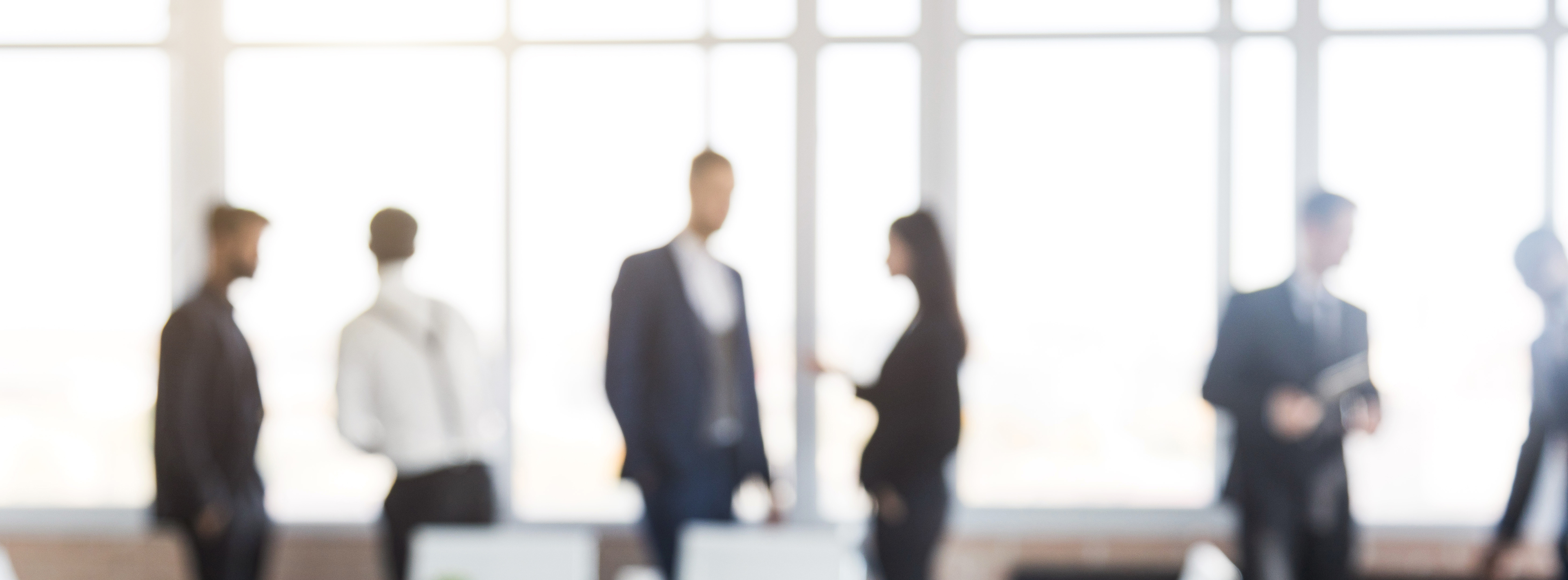 In einem modernen Büroumfeld stehen mehrere Menschen in Anzug und Businesskleidung. Sie führen Gespräche und tauschen Informationen aus, während große Fenster im Hintergrund für eine helle, offene Atmosphäre sorgen. Die Gestalten sind unscharf, was einen Fokus auf die Interaktion der Personen legt.