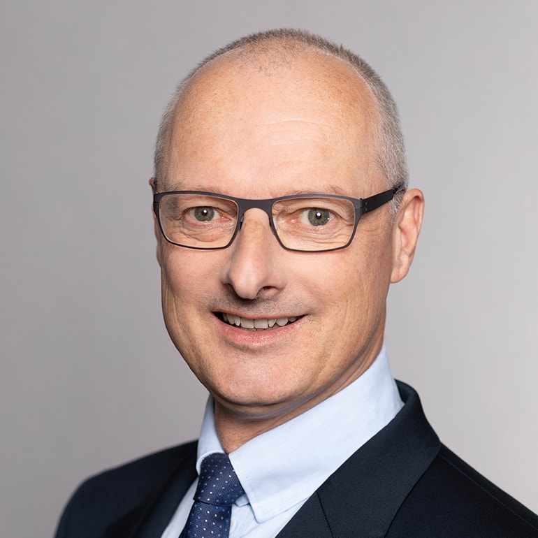 Porträt eines Mannes mit Brille, der in einem Anzug und einem hellblauen Hemd gekleidet ist. Er lächelt freundlich und hat eine glatte Kopfhaut. Der Hintergrund ist neutral und einfarbig.