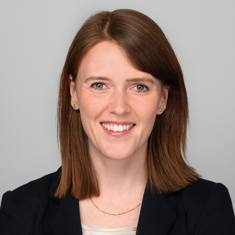 Ein Porträt einer lächelnden Frau mit braunen Haaren, die schulterlang sind. Sie trägt einen schwarzen Blazer und eine helle Bluse. Der Hintergrund ist grau. Ihr Gesicht ist freundlich und einladend.