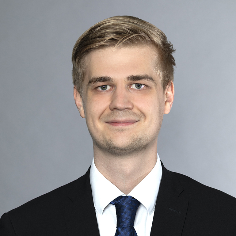 Ein junger Mann mit blonden Haaren und einem freundlichen Lächeln trägt einen schwarzen Anzug und eine blaue Krawatte. Der Hintergrund ist neutral grau.