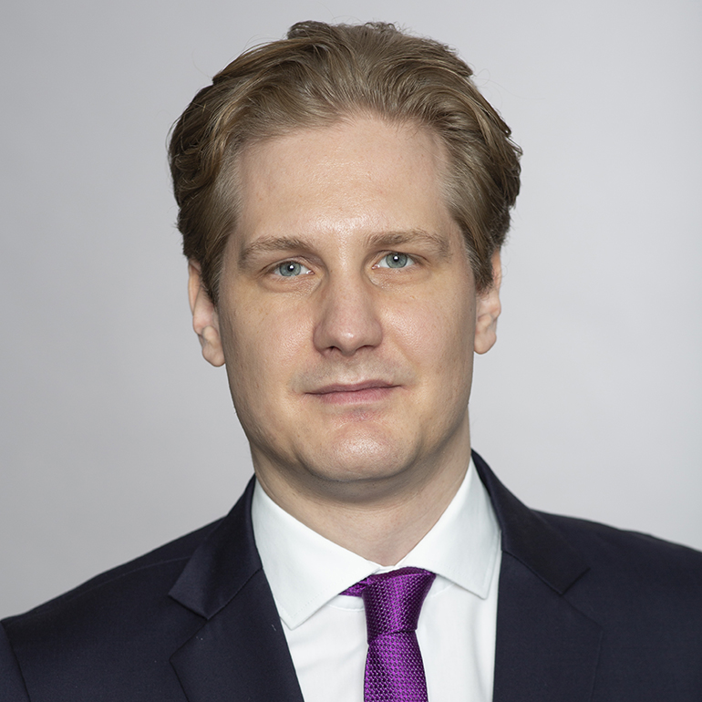 Ein lächelnder Mann mit blonden Haaren trägt einen dunklen Anzug und eine lila Krawatte. Sein Gesichtsausdruck ist freundlich und selbstbewusst. Der Hintergrund ist neutral und hell, wodurch er im Vordergrund gut zur Geltung kommt.