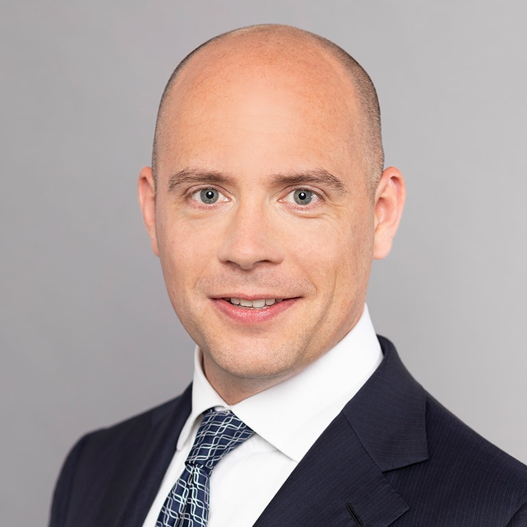 A young man with a bald head and light complexion smiles at the camera. He is dressed in a dark suit with a white shirt and a patterned tie. The background is a light gray color, keeping the focus on his face.