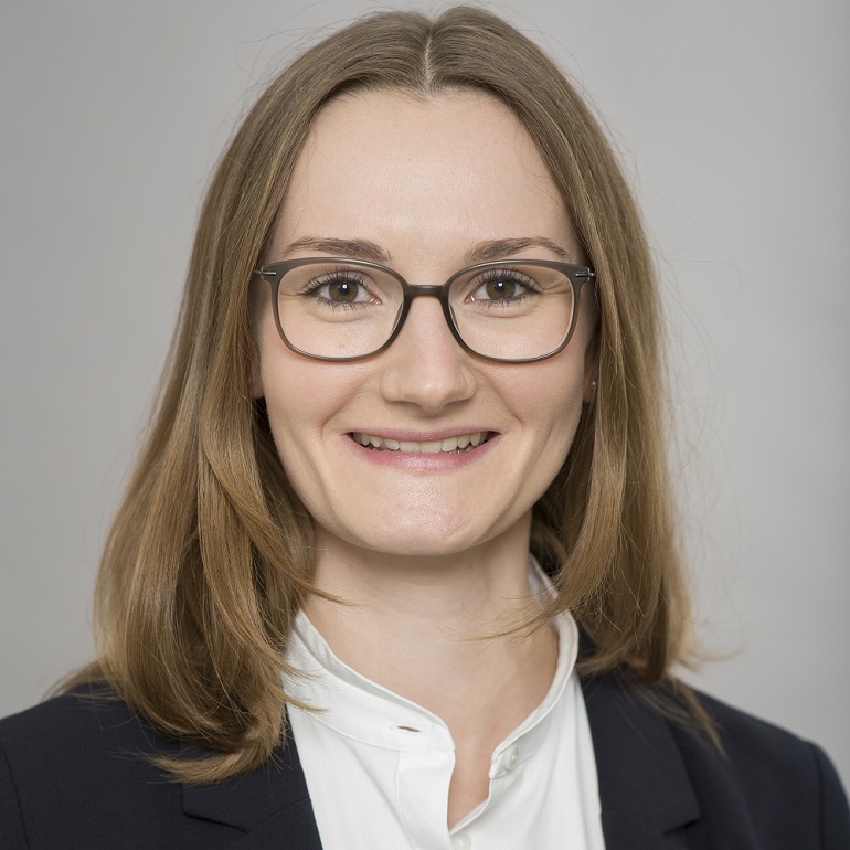 Portrait einer lächelnden Frau mit langen, braunen Haaren. Sie trägt eine schwarze Blazerjacke und ein weißes Oberteil. Die Frau hat eine Brille und steht vor einem hellen, einfarbigen Hintergrund.