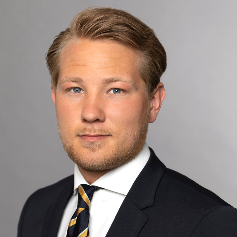 Ein junger Mann mit blonden Haaren, einem leichten Bart und blauen Augen trägt einen dunklen Anzug mit einem weißen Hemd und einer gestreiften Krawatte. Der Hintergrund ist grau und neutral gehalten. Der Blick des Mannes strahlt Selbstbewusstsein aus.