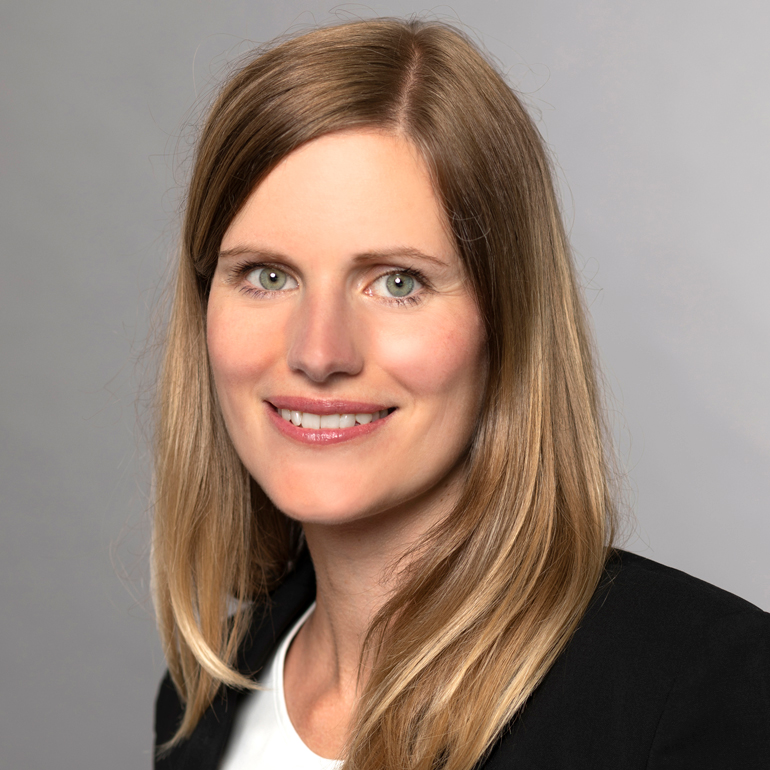 Porträt einer lächelnden Frau mit langen, blonden Haaren. Sie trägt ein schwarzes Blazer-Jackett und hat helle Haut. Der Hintergrund ist neutral und grau, wodurch der Fokus auf ihrem Gesicht liegt.