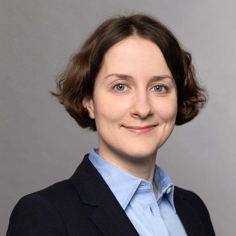 Porträt einer Frau mit kurzen, lockigen Haaren und einem freundlichen Lächeln. Sie trägt einen dunklen Blazer und ein hellblaues Hemd. Der Hintergrund ist einfarbig grau, was einen professionalen Eindruck vermittelt.