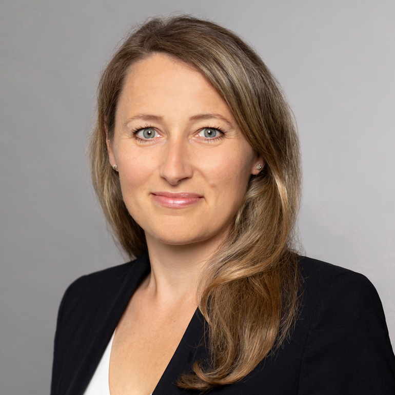 Porträt einer Frau mit langen, hellbraunen Haaren. Sie trägt eine dunkle Jacke und hat ein freundliches Lächeln. Der Hintergrund ist hellgrau, was den Fokus auf ihr Gesicht legt. Ihre blauen Augen und der natürliche Look strahlen Selbstbewusstsein aus.