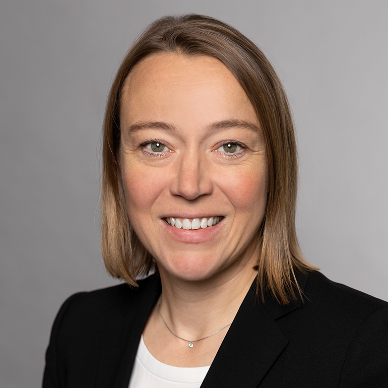 Porträtaufnahme einer lächelnden Frau mit mittellangen, braunen Haaren. Sie trägt ein schwarzes Sakko und ein weißes Oberteil. Der Hintergrund ist grau.