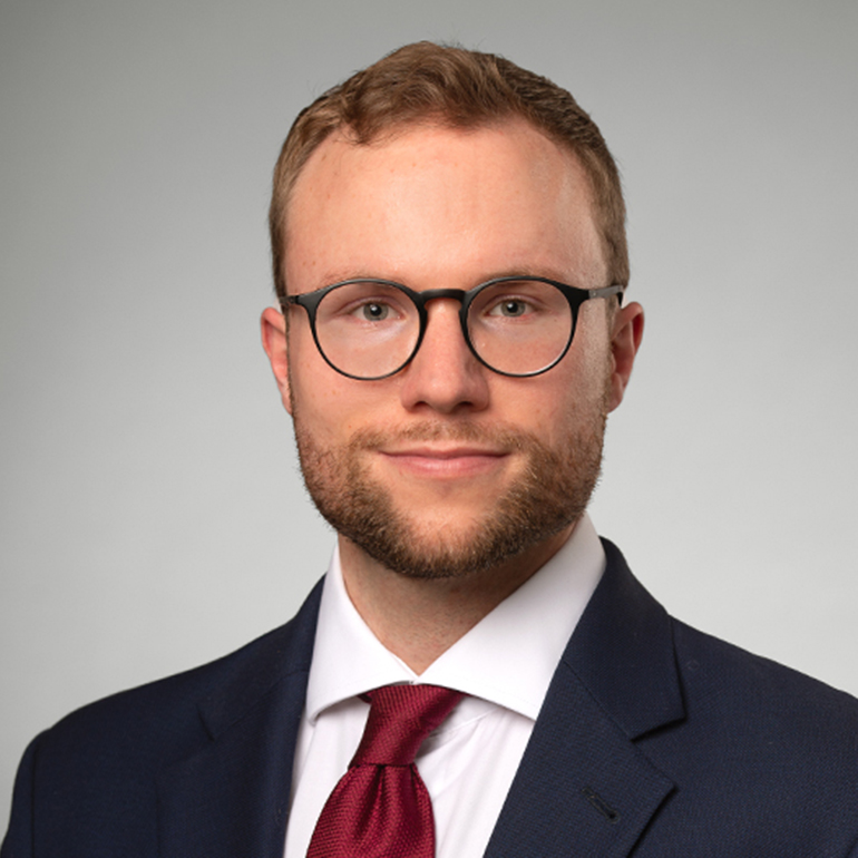 Ein junger Mann mit Brille, kurzem, blonden Haar und leichtem Bart steht vor einem einfarbigen Hintergrund. Er trägt einen dunkelblauen Anzug und ein weißes Hemd mit einer roten Krawatte. Sein Ausdruck ist freundlich und selbstbewusst.