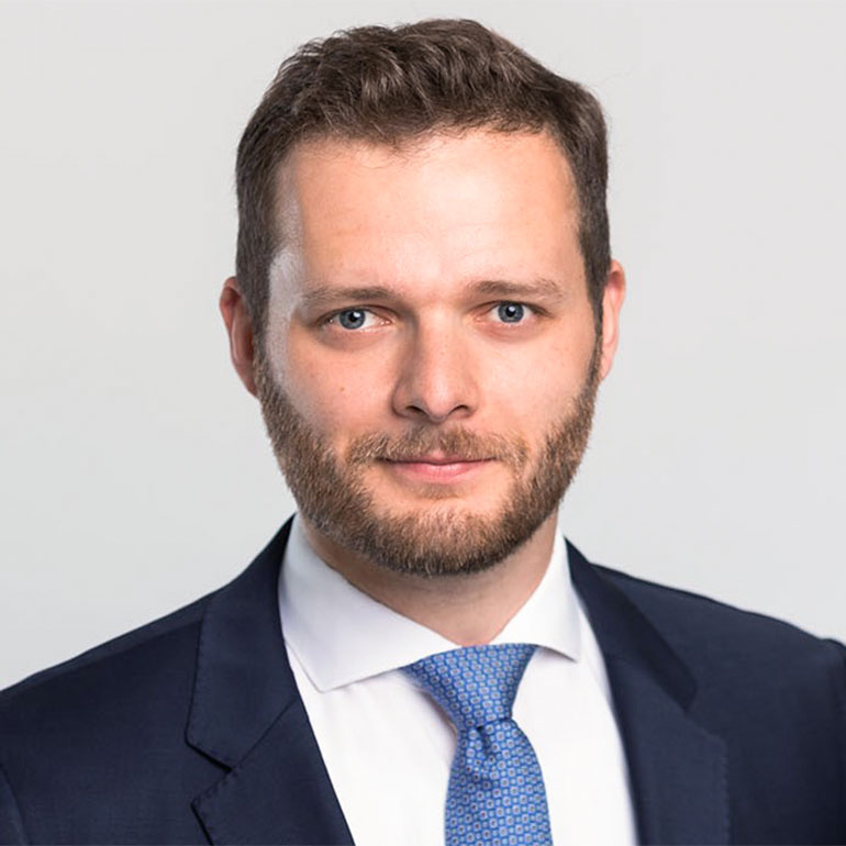 Ein Mann mit kurzem, braunem Haar und einem Bart blickt direkt in die Kamera. Er trägt einen dunklen Anzug und eine hellblaue Krawatte. Der Hintergrund ist neutral und hell. Die Gesamtstimmung wirkt professionell und freundlich.