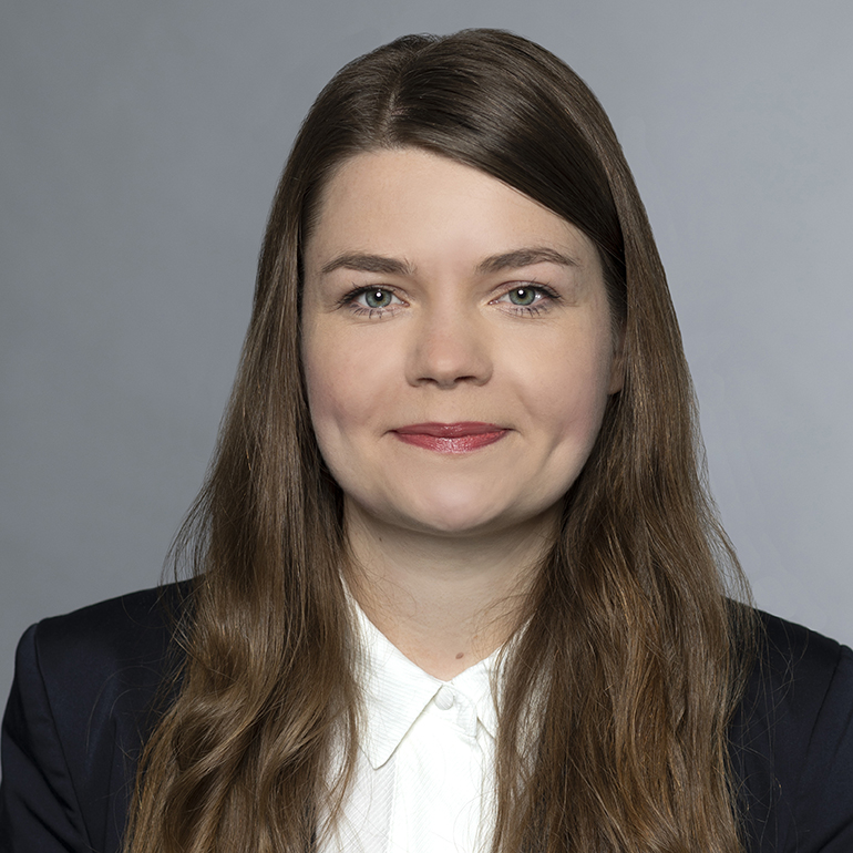 Ein Porträt einer Frau mit langen, braunen Haaren und einem freundlichen Lächeln. Sie trägt ein dunkles Blazer-Oberteil und ein helles Hemd. Der Hintergrund ist neutral und hellgrau, was den Fokus auf ihr Gesicht legt.