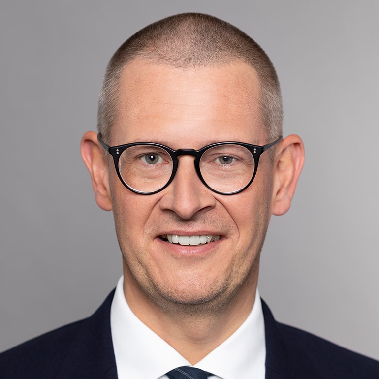 Porträt eines Mannes mit kurzen, grauen Haaren, der eine brillante Brille trägt. Er lächelt freundlich und trägt einen dunklen Anzug mit einem weißen Hemd und einer gestreiften Krawatte. Der Hintergrund ist einfarbig und neutral.