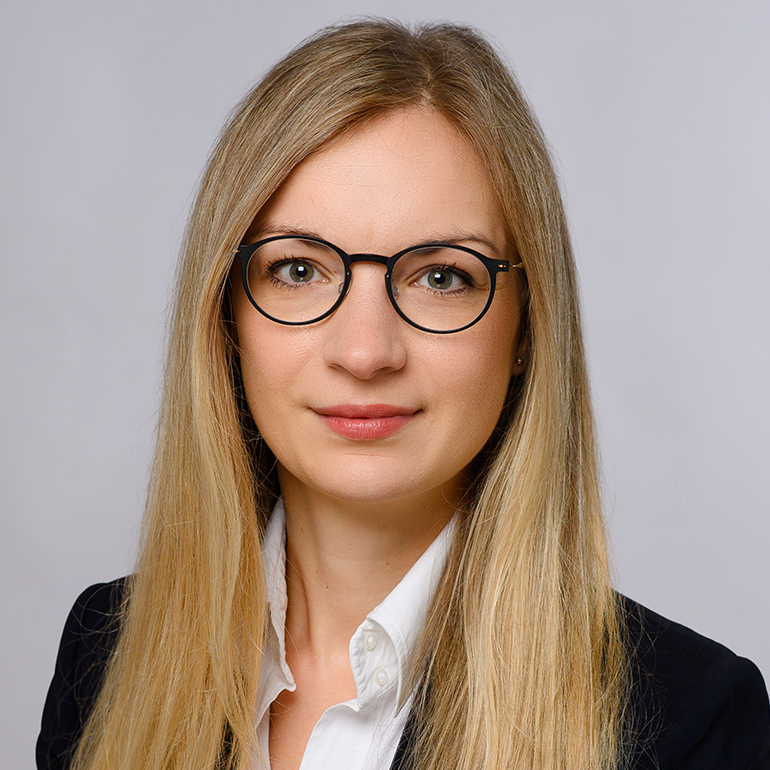 Porträt einer Frau mit langen blonden Haaren und Brille. Sie hat einen neutralen Gesichtsausdruck und trägt ein schwarzes Jackett über einem weißen Hemd. Der Hintergrund ist hell und neutral.