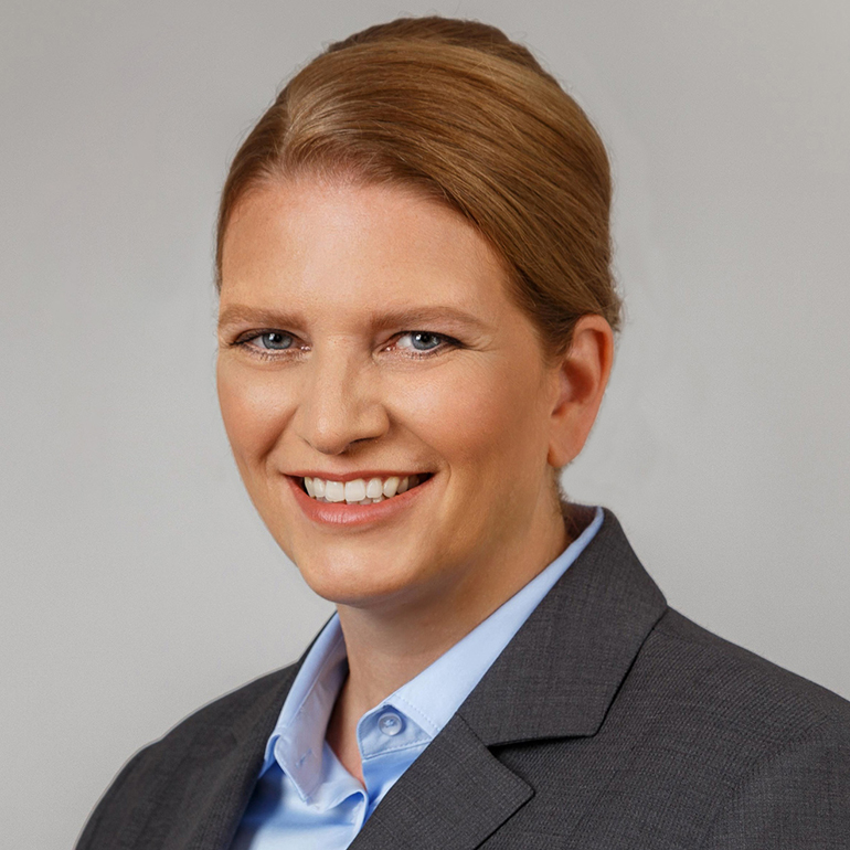 Porträt einer lächelnden Frau mit kurzen, blonden Haaren, die einen grauen Anzug und ein hellblaues Hemd trägt. Der Hintergrund ist hell und neutral, was den Fokus auf ihr freundliches Gesicht lenkt.