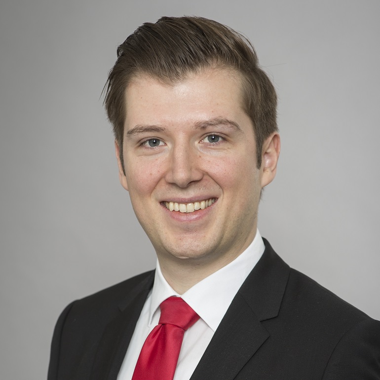 Ein junger Mann mit kurzen, braunen Haaren lächelt in die Kamera. Er trägt einen schwarzen Anzug mit einem weißen Hemd und einer roten Krawatte. Der Hintergrund ist grau und neutral gehalten.