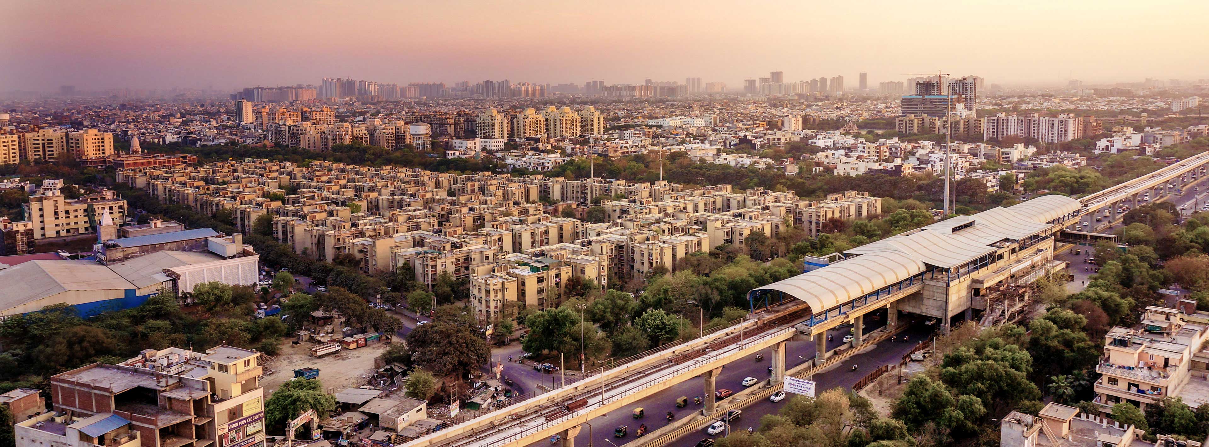 Eine weite Aussicht auf eine moderne Stadtlandschaft mit Wohngebäuden und grünen Flächen. Im Vordergrund verläuft eine Straßenbahnlinie, die zu einer Haltestelle führt. Die Skyline im Hintergrund zeigt hohe Gebäude, unter einen leicht orangefarbenen Himmel.