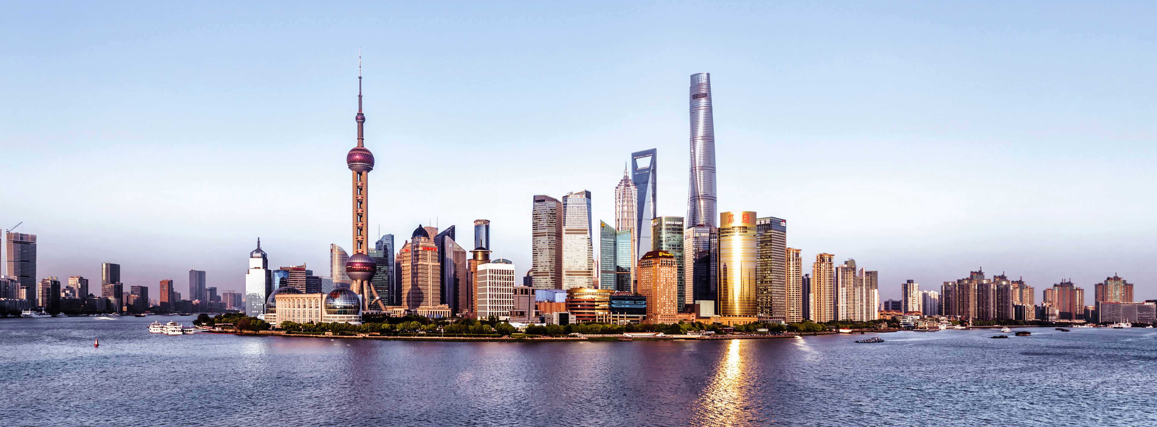 Die Skyline von Shanghai wird im Vordergrund von Hochhäusern dominiert, darunter der markante Oriental Pearl Tower und der Shanghai Tower. Das Bild zeigt den Huangpu-Fluss, der in sanften Wellen an der Stadt vorbei fließt. Der Himmel ist in sanften Farbtönen gehalten.