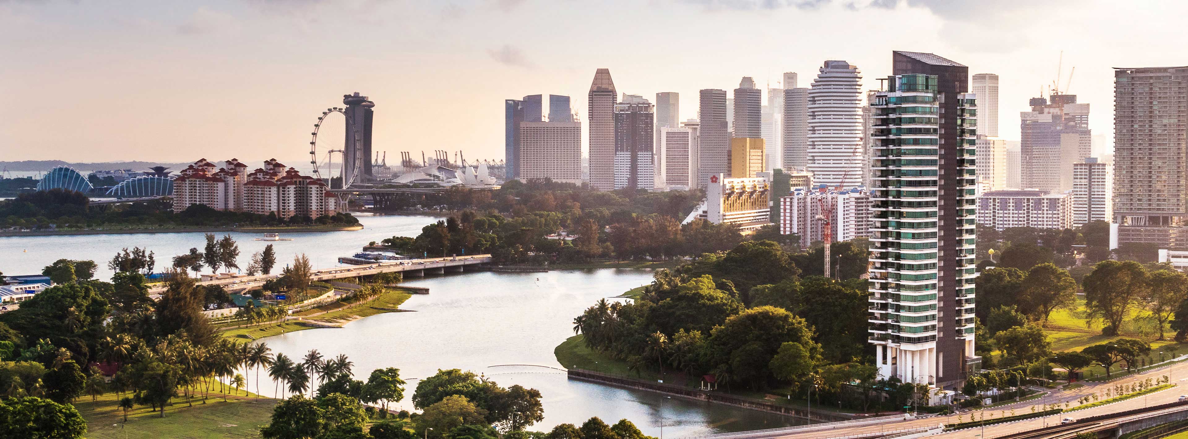 Ein Blick auf die Skyline von Singapur mit modernen Wolkenkratzern, einer grünen Uferpromenade und einem Wasserlauf. Im Hintergrund sind markante Bauwerke und ein großes Riesenrad sichtbar, während die Szene von sanften Lichtverhältnissen bei Sonnenaufgang oder Sonnenuntergang geprägt ist.