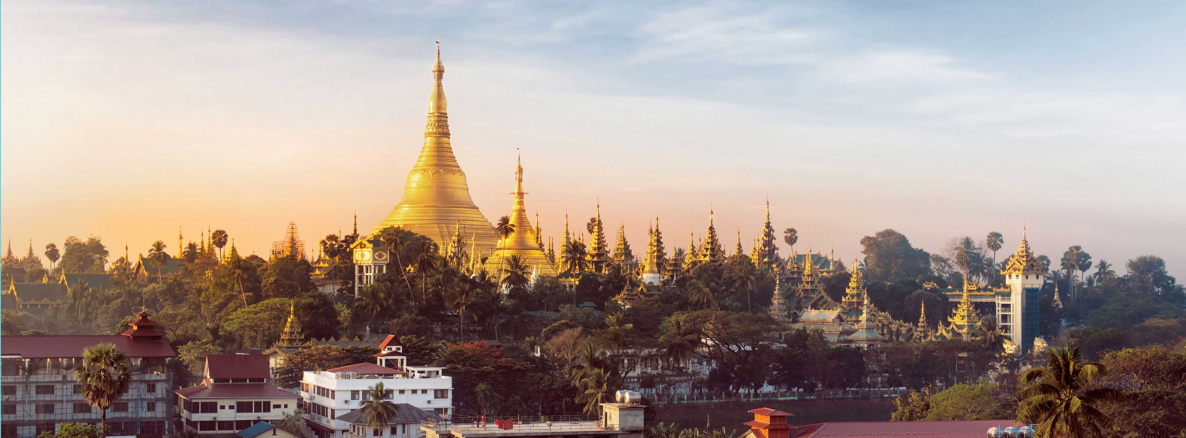 Ein atemberaubender Blick auf die goldene Shwedagon-Pagode, umgeben von weiteren Tempeln und üppigem Grün, bei Sonnenaufgang. Die Pagode erhebt sich majestätisch und spiegelt die kulturelle Bedeutung und Schönheit von Yangon, Myanmar, wider.
