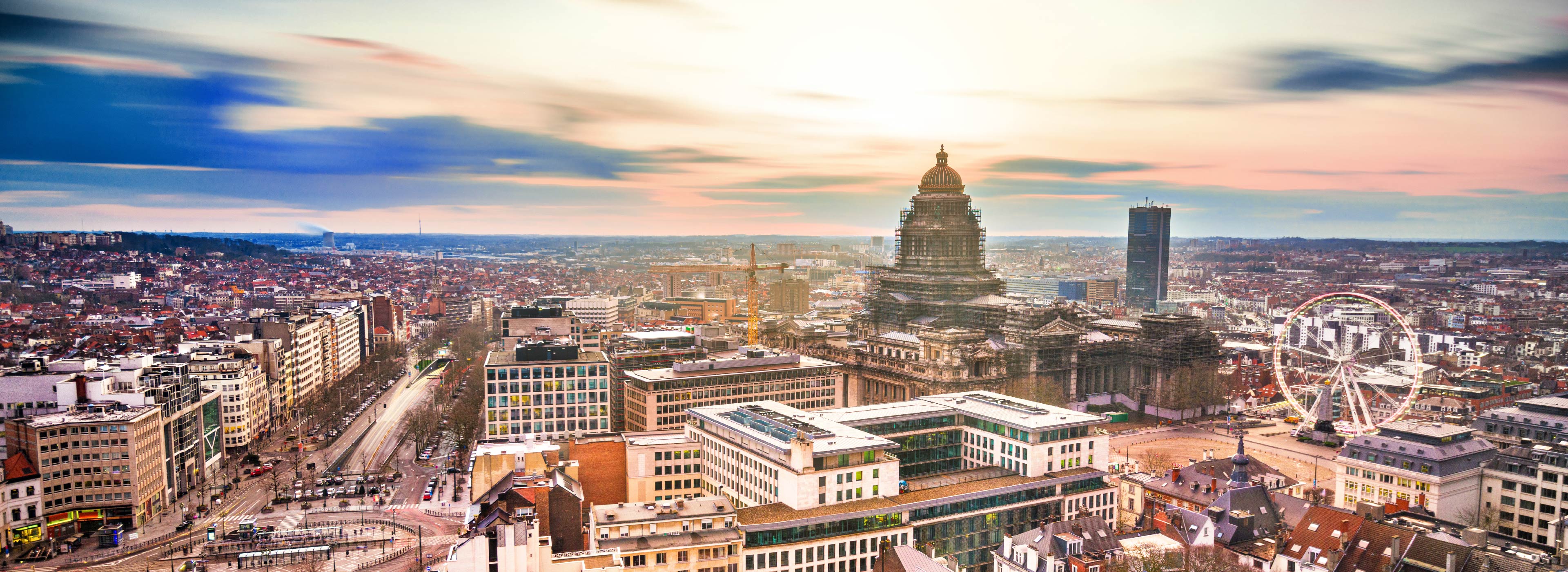 Stadtansicht bei Sonnenuntergang mit einer Mischung aus modernen Gebäuden und historischen Architekturen. Im Vordergrund sind mehrere Straßen und der große Platz zu sehen, während im Hintergrund die markante Kuppel eines historischen Gebäudes hervorsticht. Ein Riesenrad ist ebenfalls sichtbar.