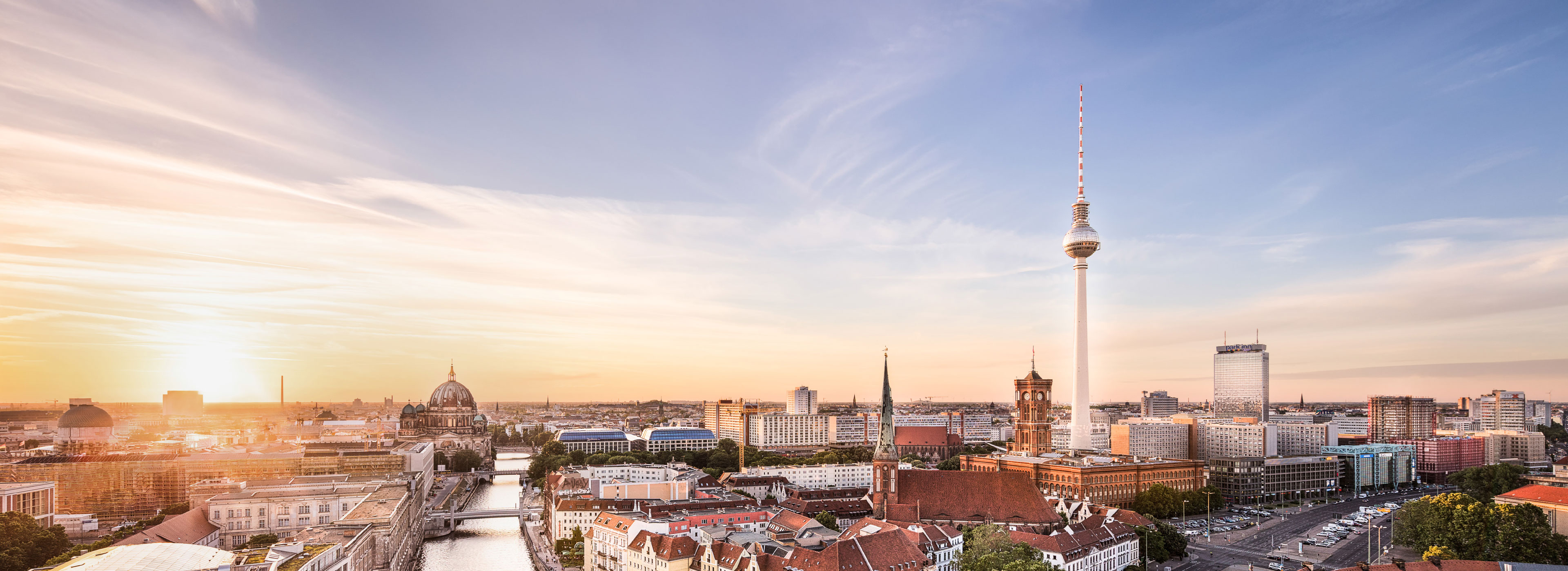 Panoramaaufnahme von Berlin bei Sonnenaufgang, die den Berliner Fernsehturm und historische Gebäude zeigt. Die ruhigere Atmosphäre der Stadt wird durch sanfte Farben des Himmels und die Reflektionen auf dem Wasser des Flusses betont.