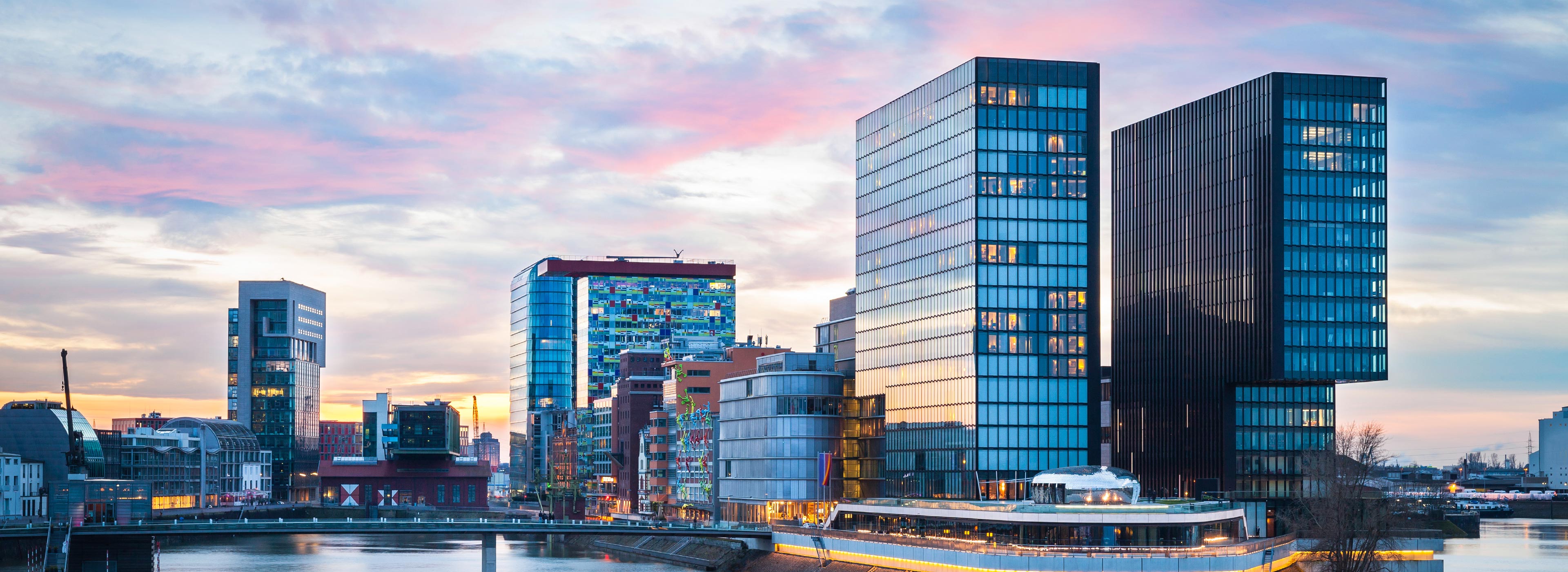 Eine Panoramaansicht einer modernen Stadtlandschaft zeigt mehrere Hochhäuser aus Glas und Stahl, die am Ufer eines Flusses stehen. Im Hintergrund sind sanfte Farbverläufe des Sonnenuntergangs zu sehen, die die Architektur der Gebäude betonen.