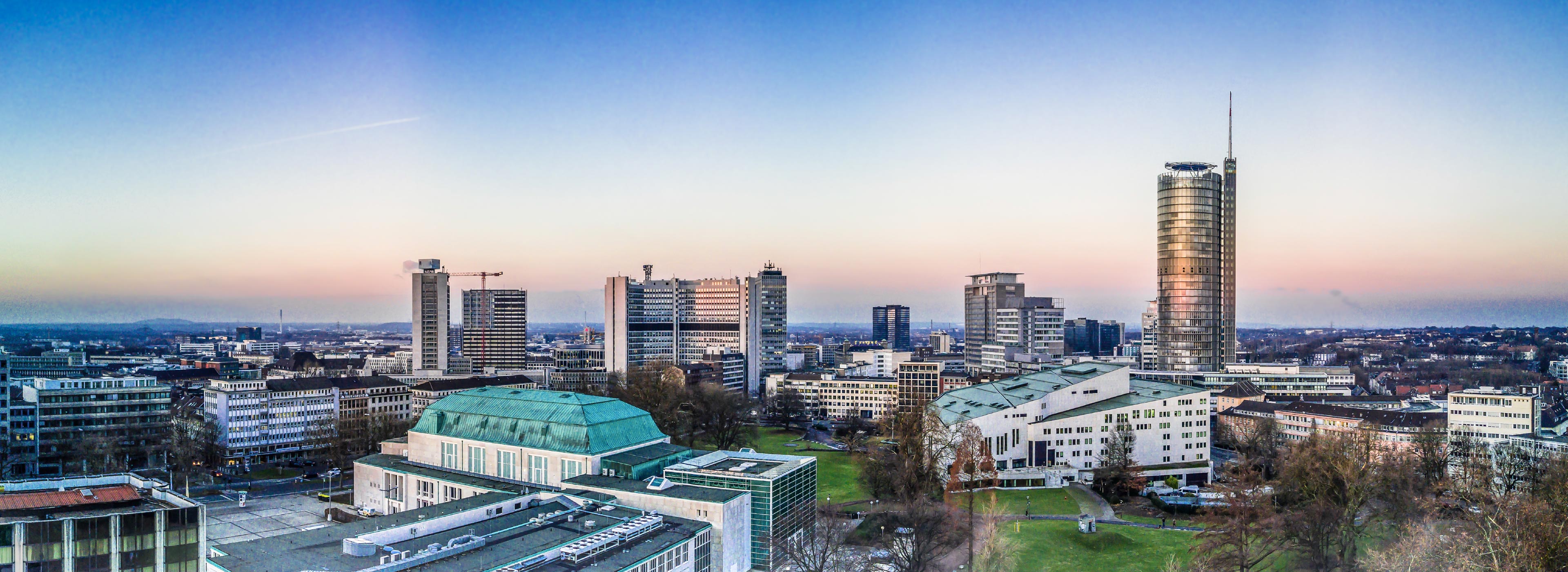 Stadtansicht bei Sonnenaufgang mit modernen Hochhäusern und einem markanten grünen Gebäude im Vordergrund. Der Himmel zeigt sanfte Farbverläufe von Blau zu Rosé, während die Stadt im Licht der Morgendämmerung erstrahlt.