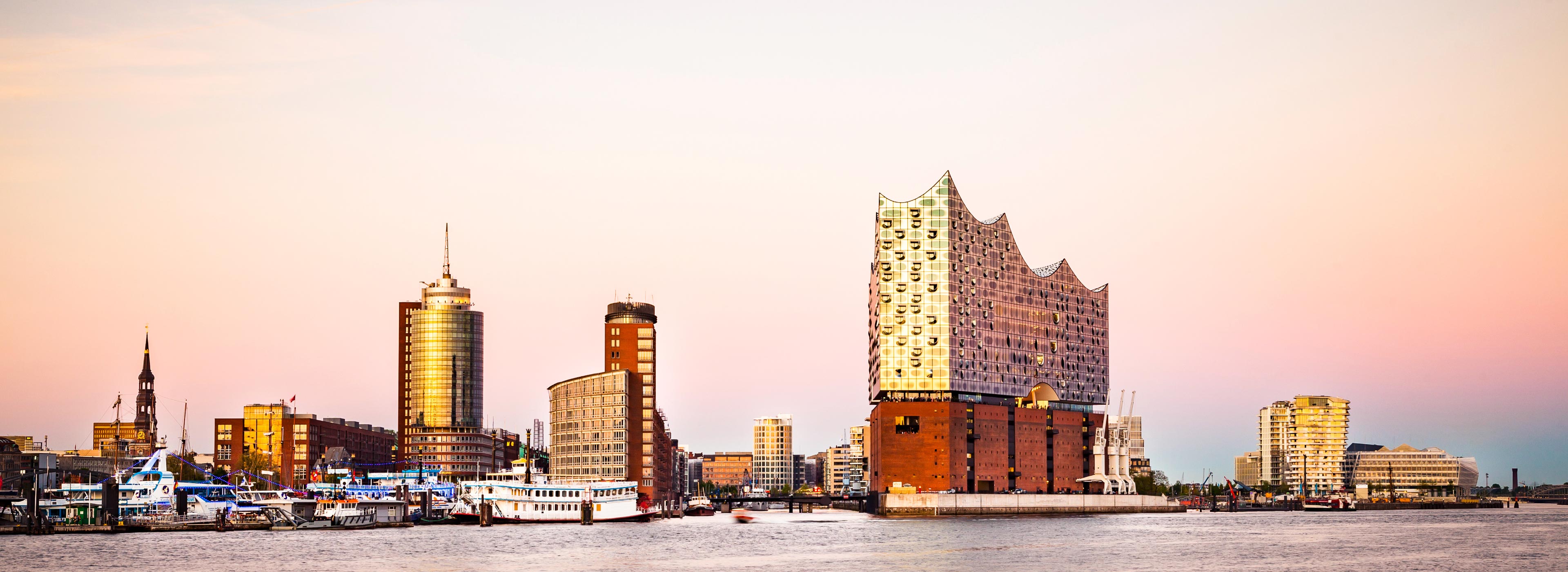 Das Bild zeigt die Hamburger Hafenlandschaft bei Sonnenuntergang. Im Vordergrund ist die Elbphilharmonie zu sehen, ein modernes, wellenförmiges Gebäude. Umgeben ist es von verschiedenen anderen Gebäuden und Booten am Wasser, während der Himmel in sanften Pastellfarben leuchtet.