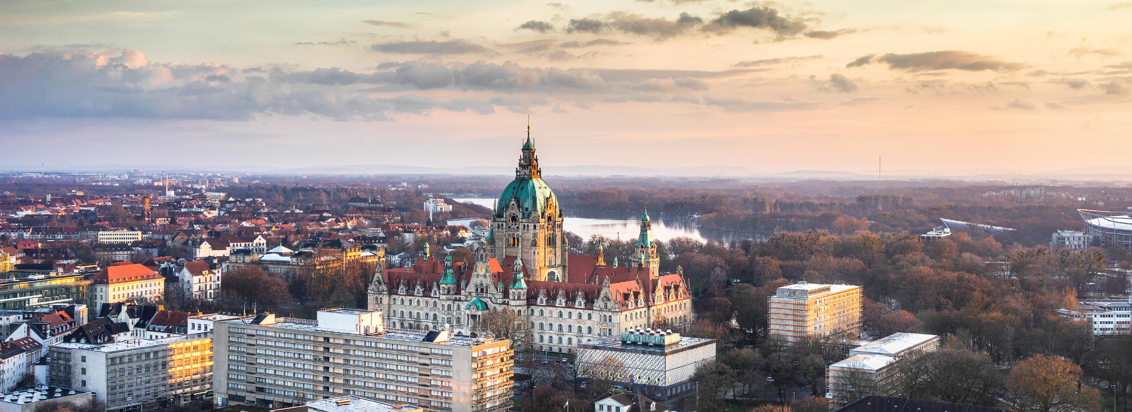 Ein Panoramablick auf Hannover bei Sonnenuntergang. Im Mittelpunkt steht das prächtige Rathaus mit seiner auffälligen Kuppel, umgeben von modernen und historischen Gebäuden. Der Himmel zeigt angenehme Farbtöne, während die Stadt im warmen Licht erstrahlt.