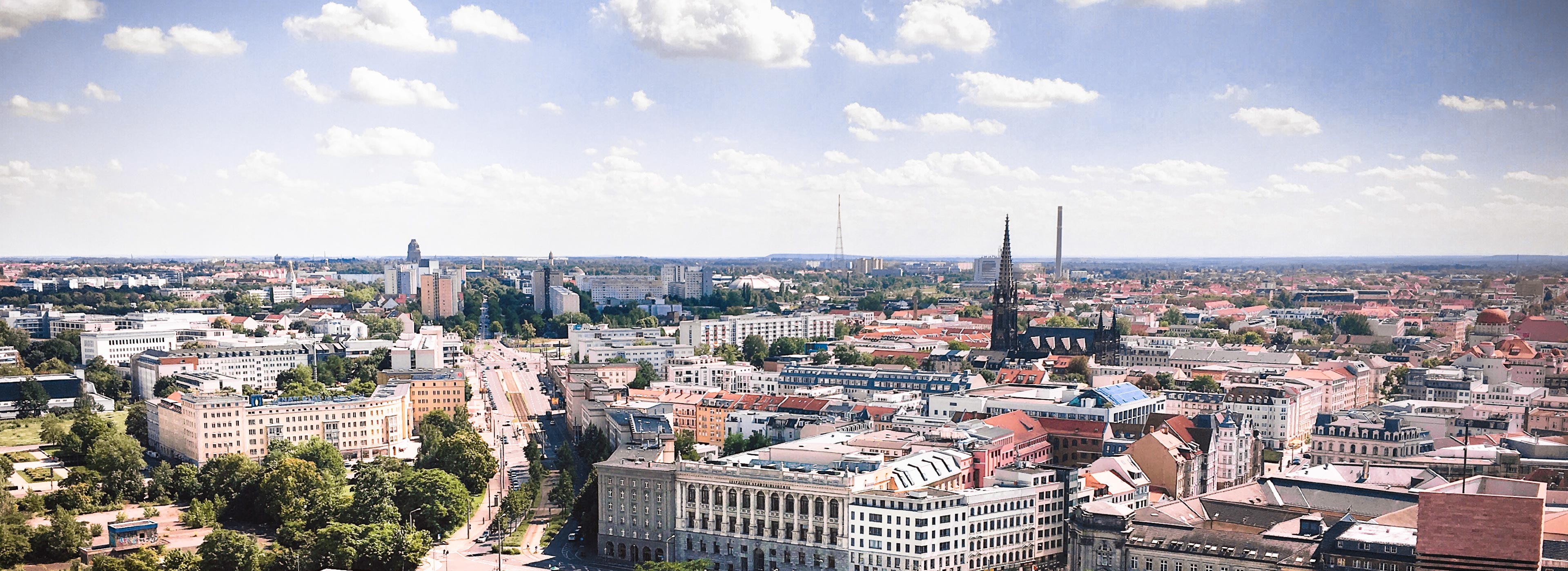 Panoramablick auf eine Stadtlandschaft mit modernen Gebäuden und einigen historischen Kirchen. Die Skyline wird von hohen Türmen und einer grünen Umgebung begleitet. Ein klarer Himmel mit wenigen Wolken rundet das Bild ab.