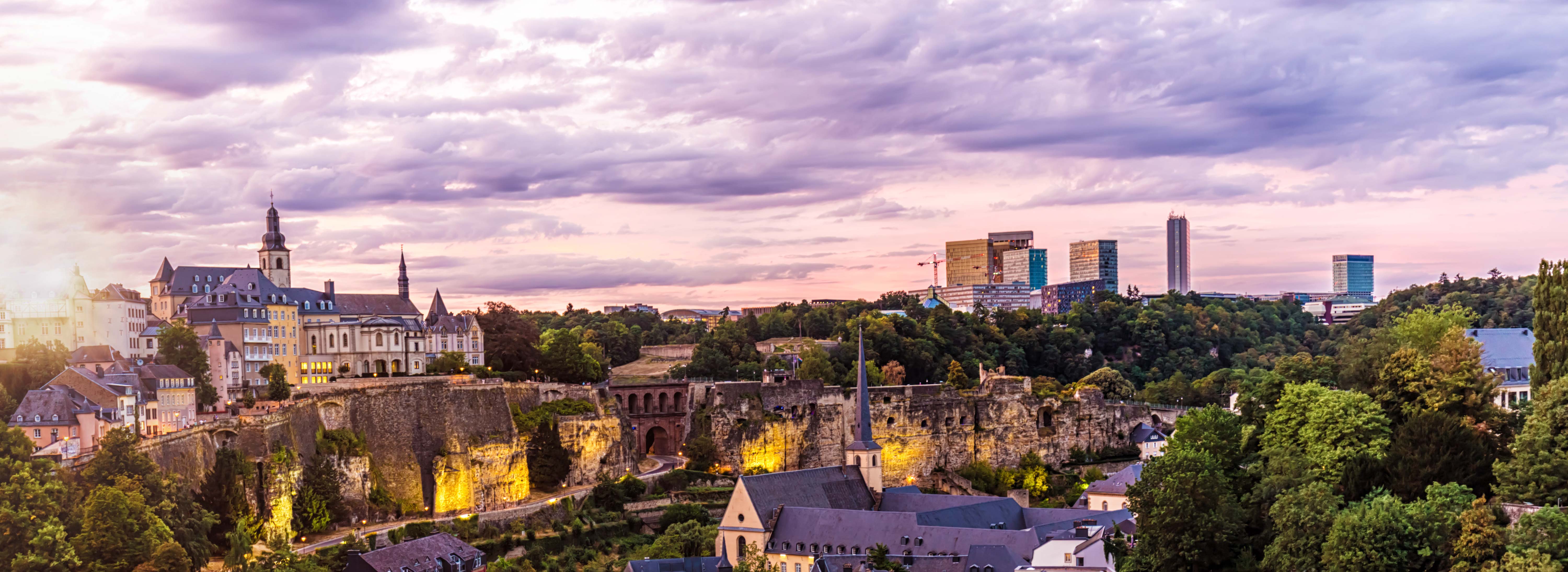 Panoramablick auf die Stadt Luxemburg bei Sonnenuntergang. Im Vordergrund historische Gebäude und eine befestigte Stadtmauer. Im Hintergrund moderne Wolkenkratzer, die das Stadtbild ergänzen. Sanfte Farben des Himmels und des Lichts schaffen eine stimmungsvolle Atmosphäre.