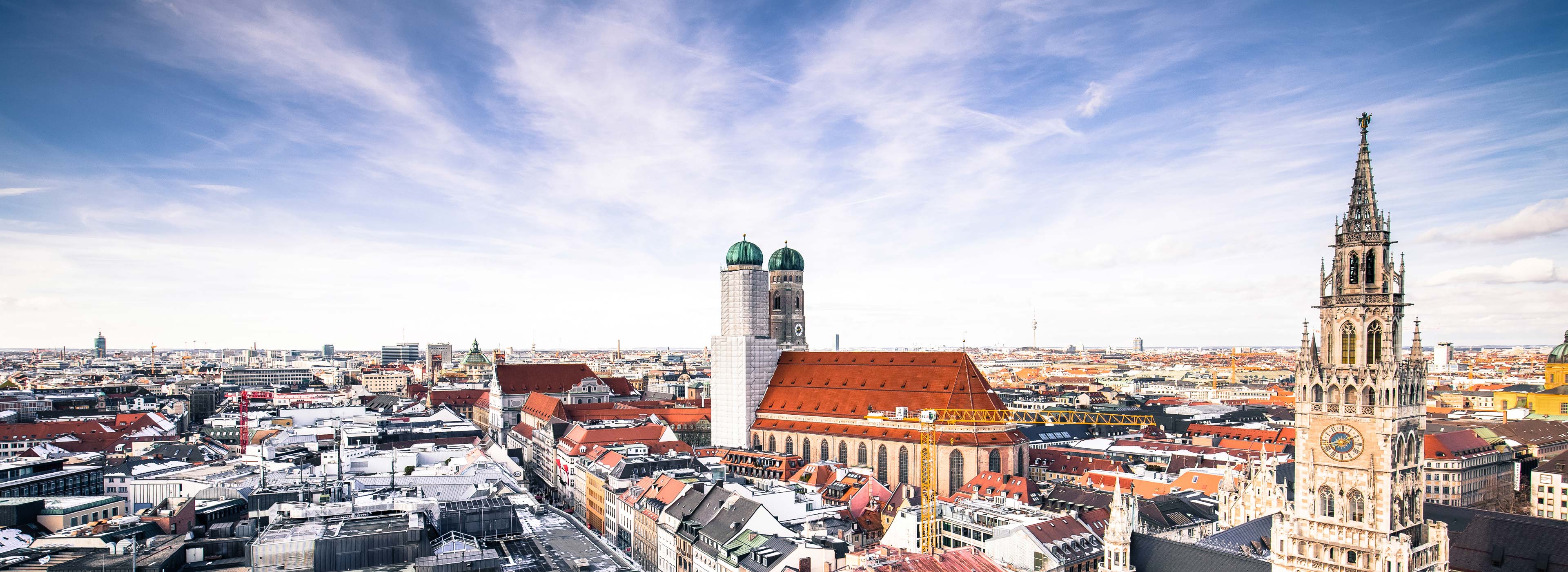 Blick auf die Skyline von München mit markanten Gebäuden wie der Frauenkirche und dem Alten Peter. Der Himmel ist blau mit einigen Wolken. Die Stadtlandschaft erstreckt sich weit und zeigt eine Mischung aus historischen und modernen Architektur.