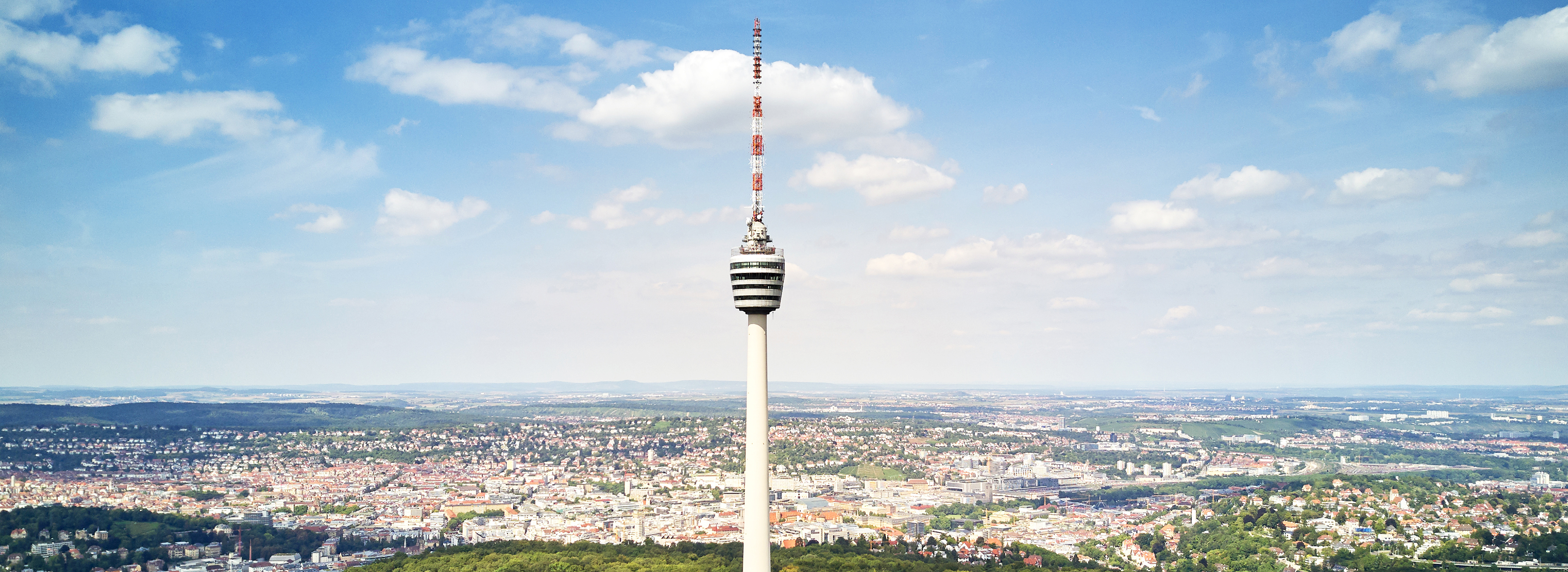 Eine Luftaufnahme eines hohen Fernsehturms, umgeben von einer Stadtlandschaft. Der Turm hat eine markante, zylindrische Form mit einer rot-weißen Antenne an der Spitze. Im Hintergrund sind weitläufige grüne Hügel und ein größeres Stadtgebiet unter einem hellblauen Himmel mit einigen Wolken sichtbar.