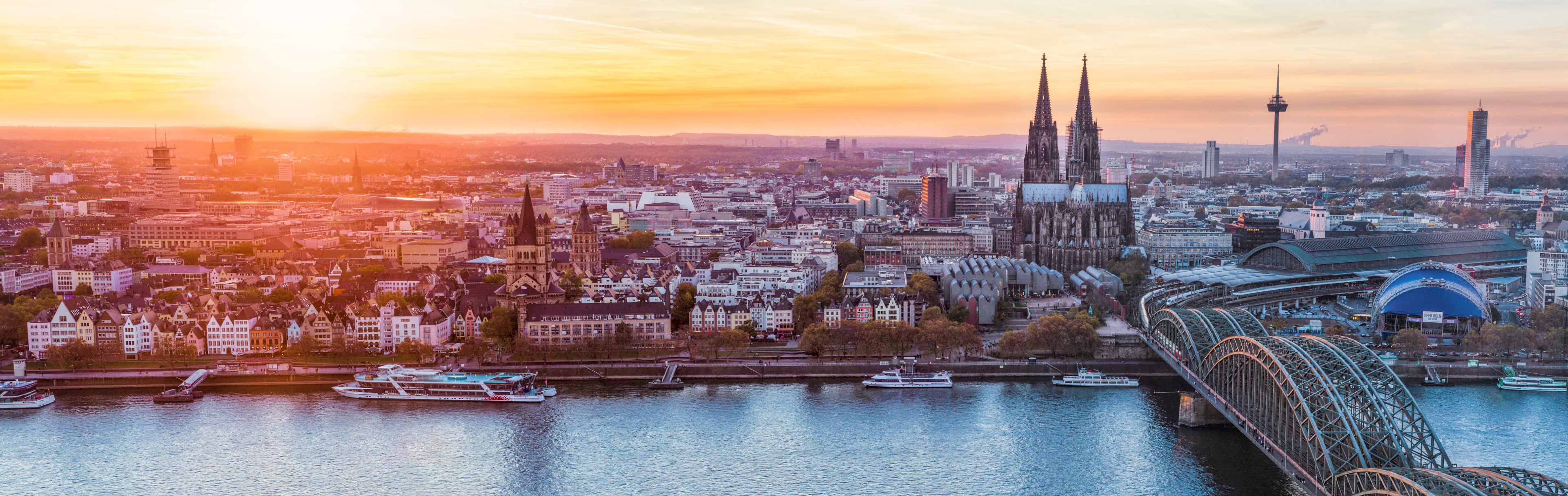 Ein Panoramablick auf Köln während des Sonnenuntergangs. Im Vordergrund ist der Rhein zu sehen, mit Booten, die darauf fahren. Im Hintergrund ragt der Kölner Dom mit seinen markanten Türmen auf, umgeben von der Skyline der Stadt und sanften Hügeln in der Ferne.