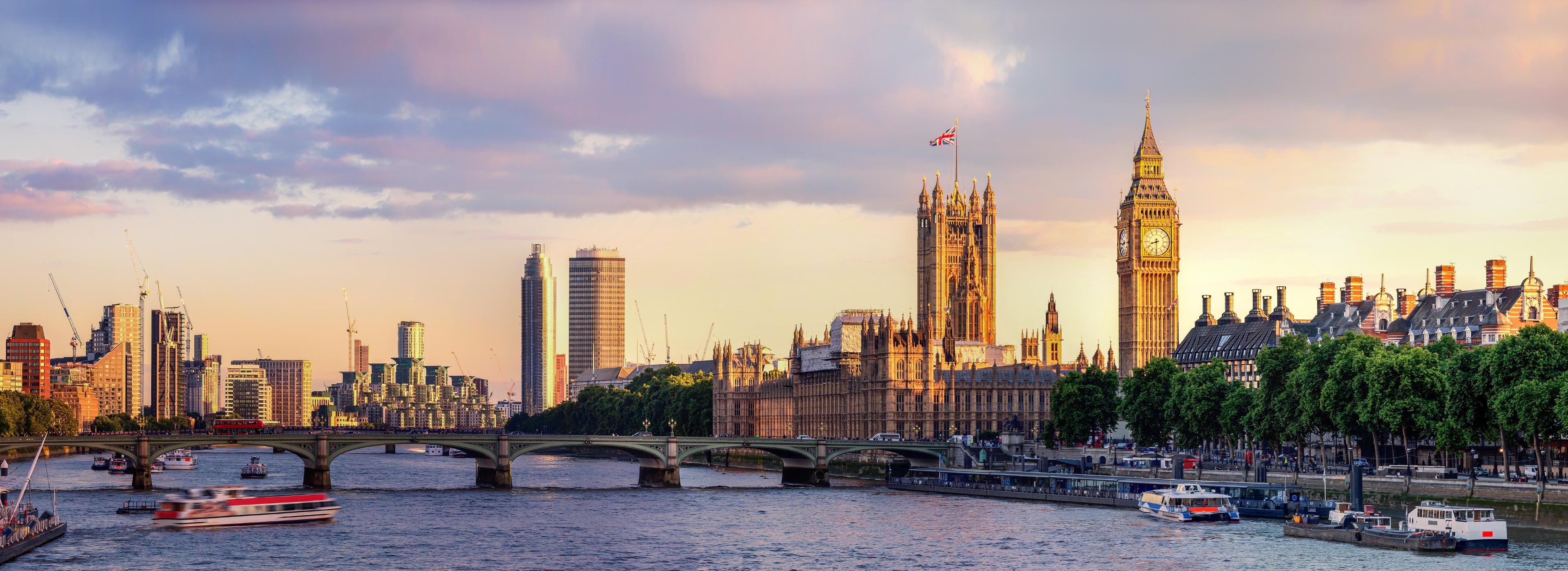 Panoramaansicht von London bei Sonnenuntergang. Im Vordergrund der Fluss Thames mit Booten, im Hintergrund der Big Ben und das Parlamentsgebäude, umgeben von modernen Wolkenkratzern. Die warme Abendhimmelfärbung verleiht der Szene eine einladende Atmosphäre.