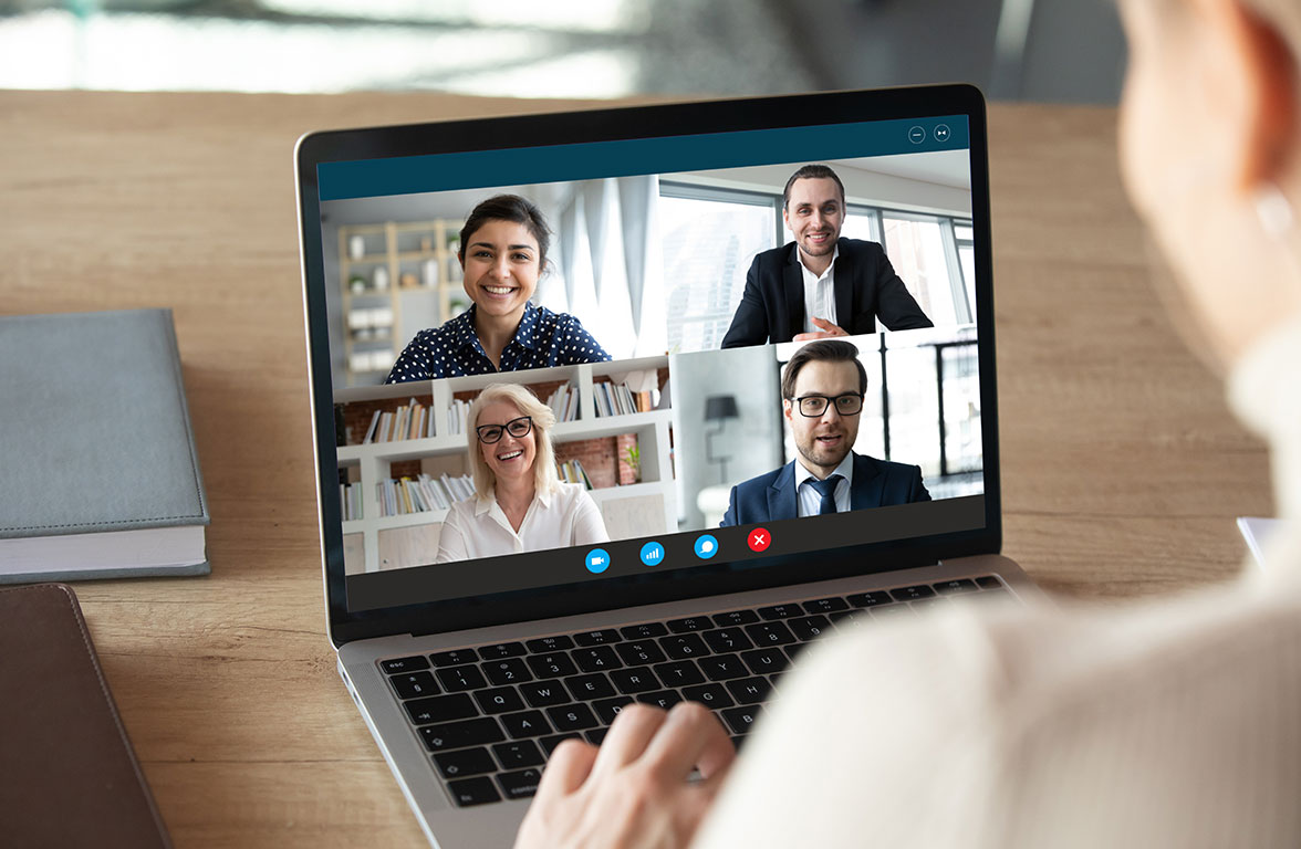 Eine Person sitzt vor einem Laptop, auf dem ein Videokonferenz-Bildschirm mit vier Teilnehmern zu sehen ist. Die Teilnehmer lächeln freundlich und kommunizieren aktiv. Der Hintergrund zeigt Bücherregale und Fenster, die Tageslicht hereinlassen.