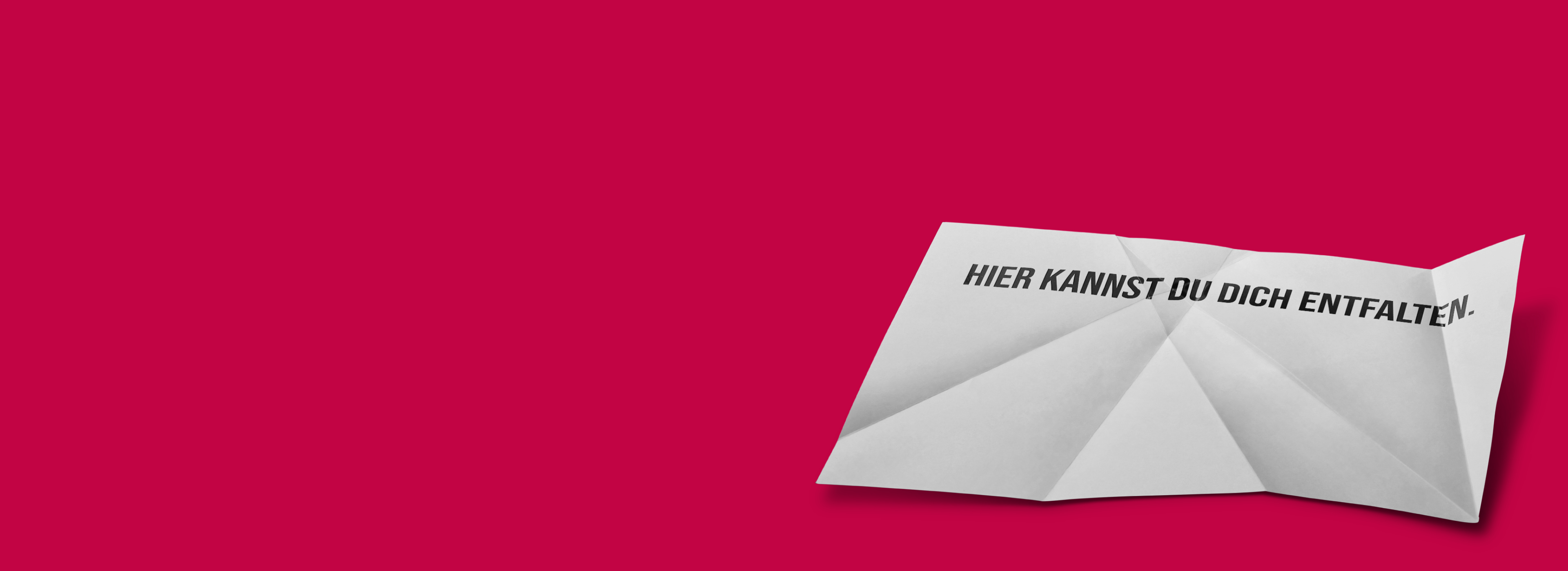 Ein zusammengefaltetes, weißes Blatt Papier mit der Aufschrift „HIER KANNST DU DICH ENTFALTEN.“ auf einem roten Hintergrund. Das Bild vermittelt eine Botschaft der Entfaltung und Möglichkeiten.