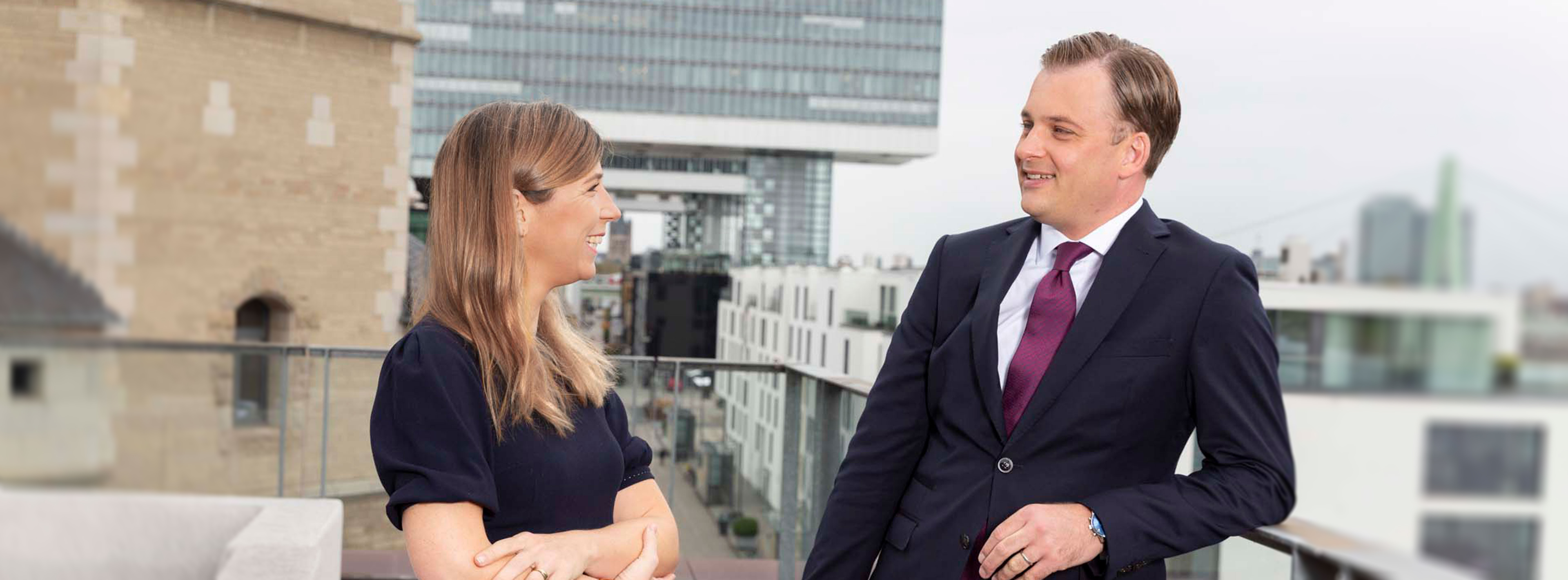 Zwei beruflich gekleidete Personen stehen auf einer Terrasse und unterhalten sich. Die Frau trägt ein dunkles Kleid, der Mann einen Anzug mit Krawatte. Im Hintergrund sind moderne Gebäude und eine Brücke zu sehen, die eine städtische Umgebung darstellt.