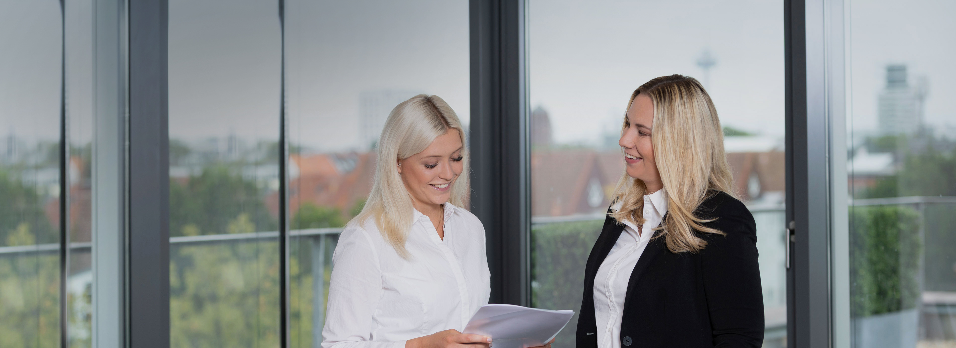 Zwei Frauen stehen in einem modernen Büroumfeld und unterhalten sich. Eine Frau trägt ein weißes Hemd und hält Papiere in der Hand, während die andere eine schwarze Jacke trägt. Beide lächeln und wirken freundlich. Im Hintergrund sind große Fenster mit Blick auf die Stadt.