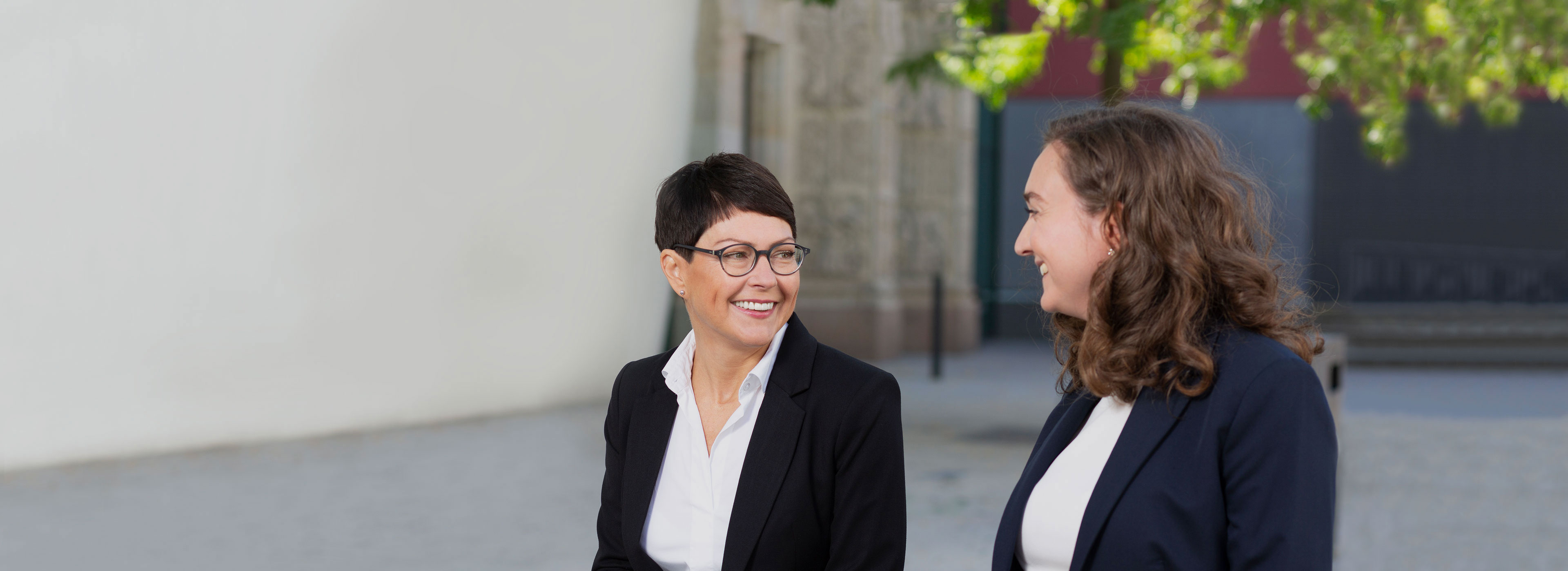 Zwei Frauen in Anzügen sitzen zusammen und lachen. Sie wirken freundlich und unterhalten sich angeregt. Im Hintergrund sind unscharfe Gebäudeteile und ein Baum sichtbar.