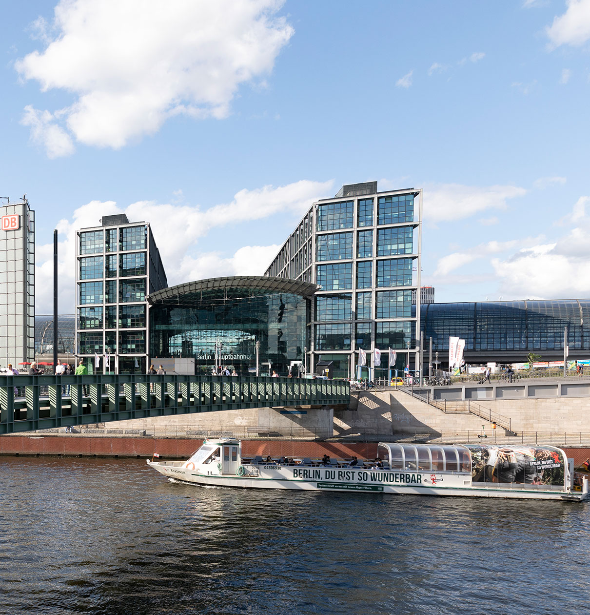 Bildbeschreibung: Die moderne Architektur des Berliner Hauptbahnhofs ist prominent sichtbar, mit großen Glasflächen und mehreren Etagen. Im Vordergrund fährt ein Schiff auf dem Wasser, auf dessen Seite der Schriftzug „Berlin, du bist so wunderbar“ zu lesen ist.