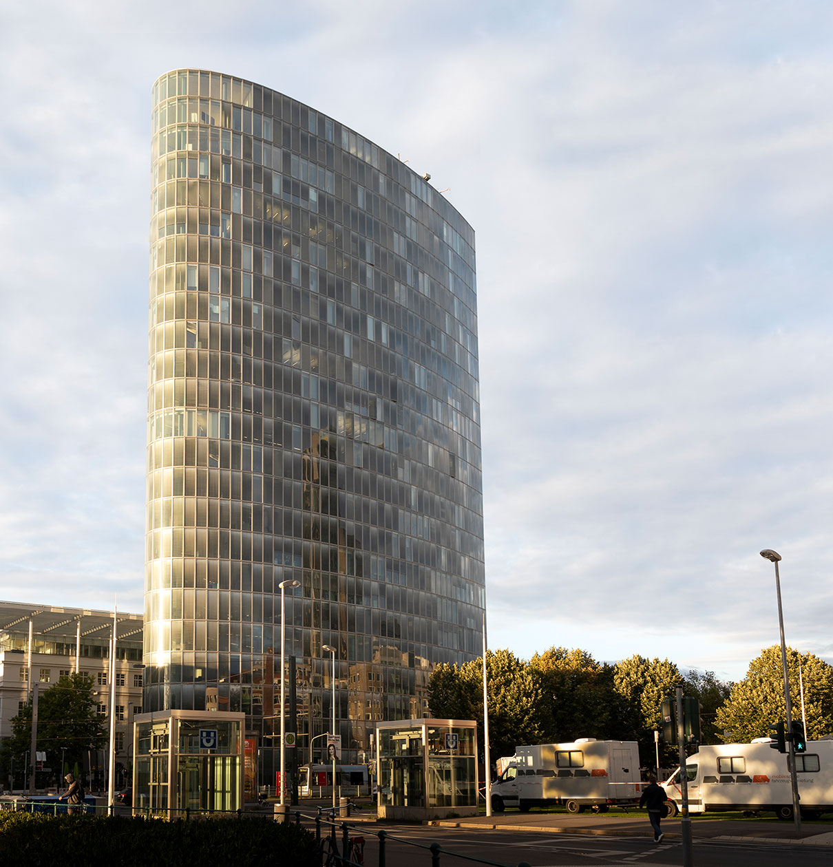 Ein modernes, hoch aufragendes Gebäude mit einer gläsernen Fassade, die das Licht reflektiert. Im Vordergrund stehen einige Bushaltestellen und graue Lieferfahrzeuge. Der Himmel ist teilweise bewölkt, und um das Gebäude herum sind Bäume zu sehen.