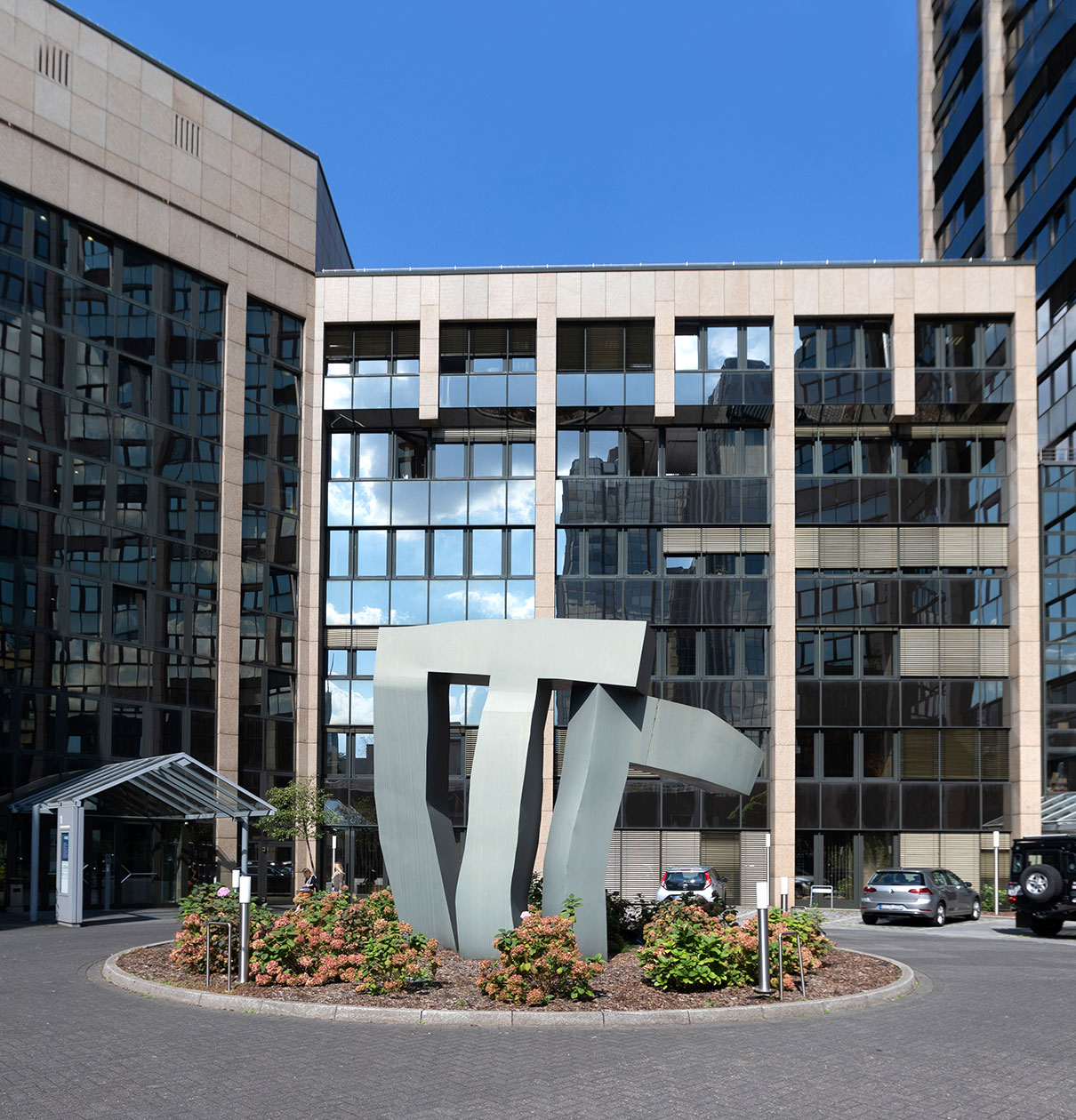 Ein moderner Gebäude- und Kunstwerkkomplex. Im Vordergrund steht eine große, abstrakte Skulptur in Grau. Die Rückseite zeigt mehrere Glasfenster eines mehrstöckigen Gebäudes, das Himmel und Wolken reflektiert. Der Eingang ist durch ein Glasdach geschützt.