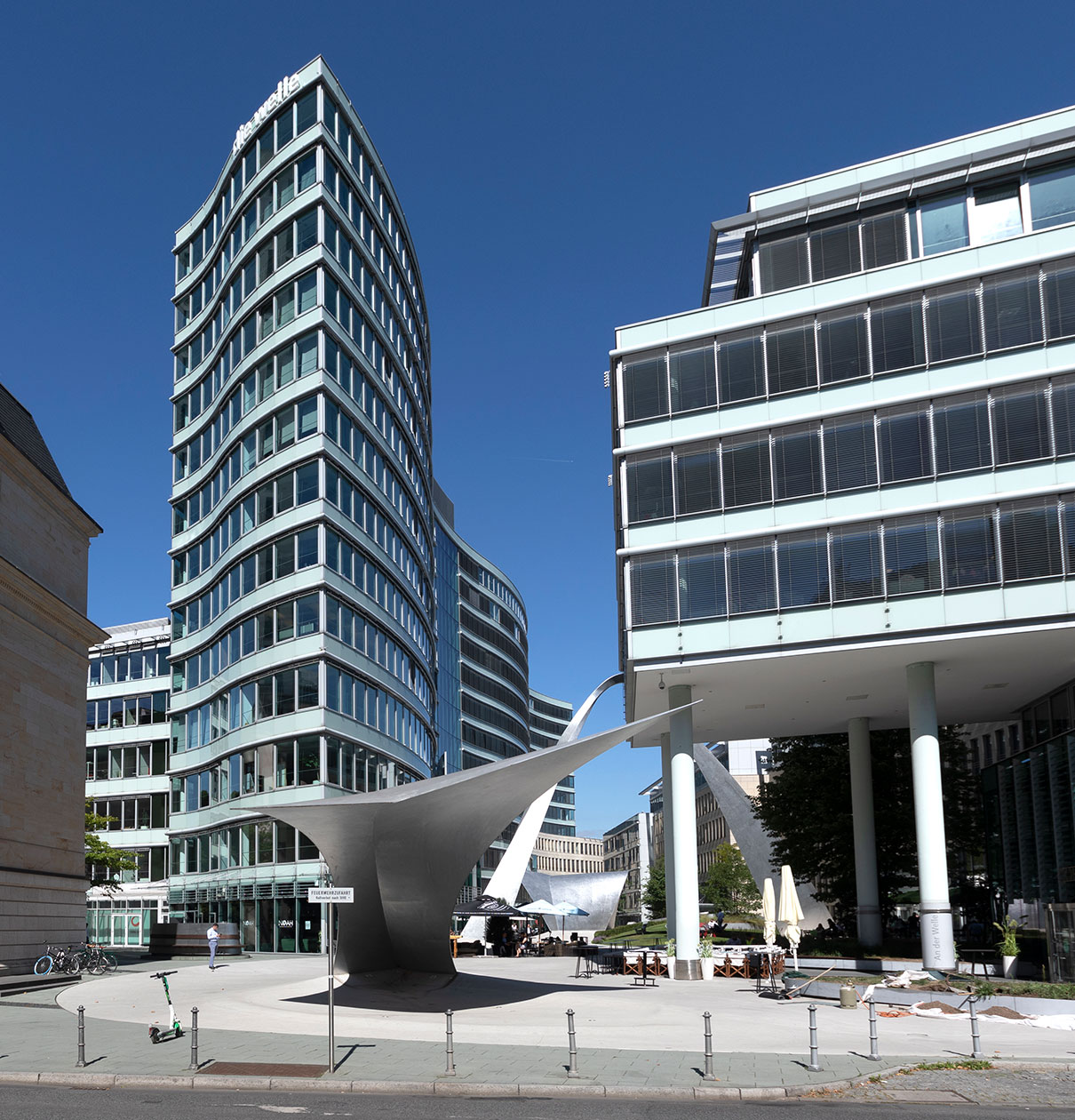 Moderne Architektur in einer Stadtlandschaft: Ein geschwungenes Gebäude mit großen Fenstern steht neben einem weiteren Glasgebäude. Im Vordergrund befindet sich eine auffällige, skulpturale Struktur, auf einem Platz mit einigen Tischen und Stühlen. Der Himmel ist klar und blau.