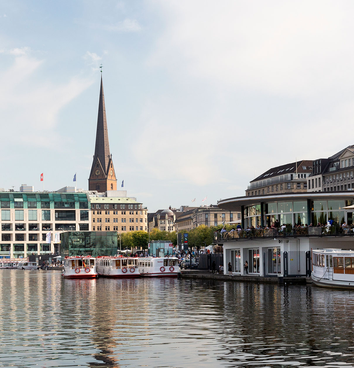 Eine Ansicht eines Gewässers mit mehreren Booten im Vordergrund. Im Hintergrund steht ein markanter Kirchturm mit einer spitzen Spitze. Auf der linken Seite sind moderne Gebäude, während rechts ein Restaurant mit Gästen auf der Terrasse zu sehen ist. Der Himmel ist hell und bewölkt.