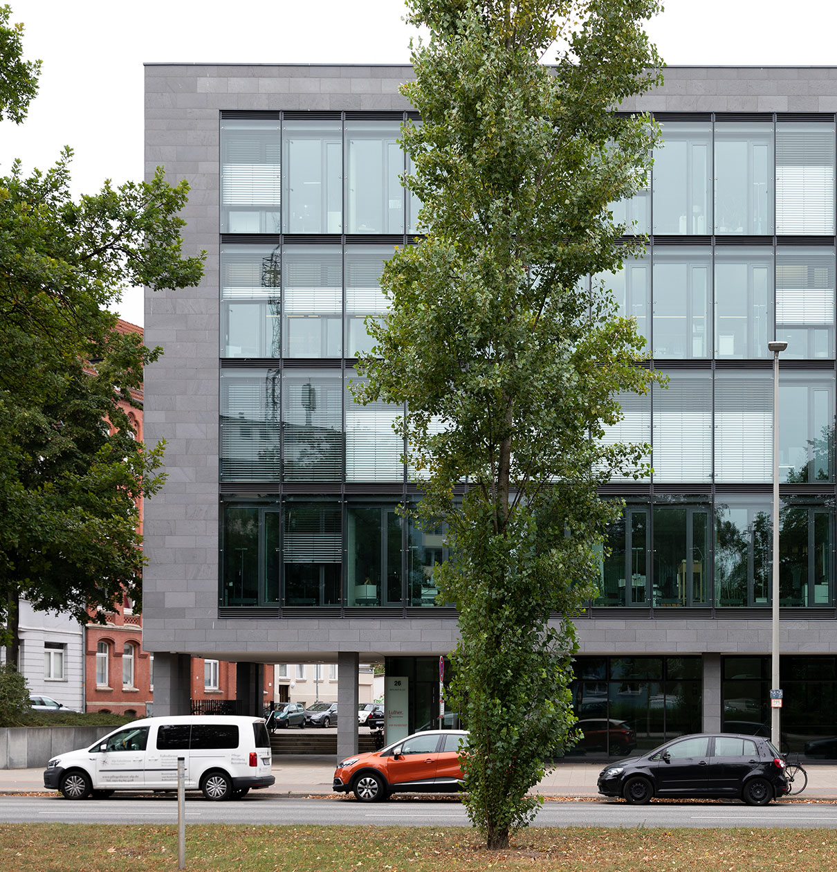 Ein modernes, mehrstöckiges Gebäude mit großen Fenstern ist im Vordergrund zu sehen. Vor dem Gebäude stehen mehrere Fahrzeuge, darunter ein weißer und ein orangefarbener Wagen. Im Vordergrund wächst ein Baum, der teilweise das Gebäude verdeckt.