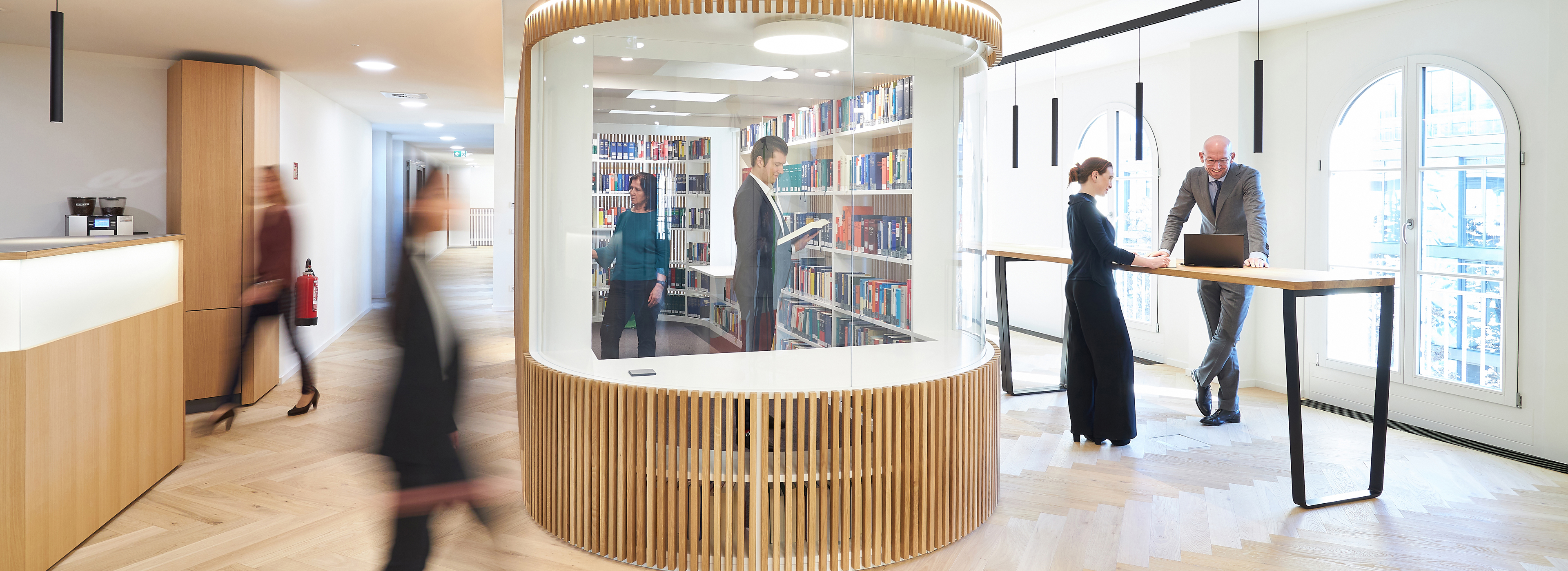 In einer modernen Büroumgebung stehen mehrere Personen zusammen. Im Hintergrund ist eine Bibliothek mit Regalen voller Bücher sichtbar. Eine Person sitzt an einem Tisch mit einem Laptop, während andere in einem Gespräch vertieft sind. Das Gesamtbild vermittelt eine produktive Atmosphäre.