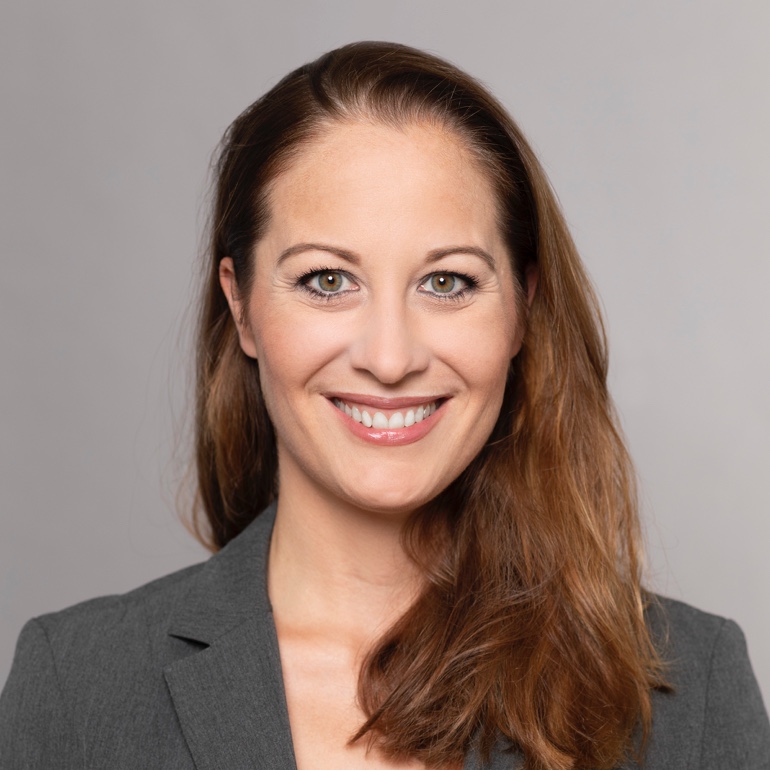 Porträt einer lächelnden Frau mit langen, braunen Haaren. Sie trägt einen grauen Blazer und hat einen freundlichen Ausdruck. Der Hintergrund ist neutral und hell, was den Fokus auf ihr Gesicht lenkt.